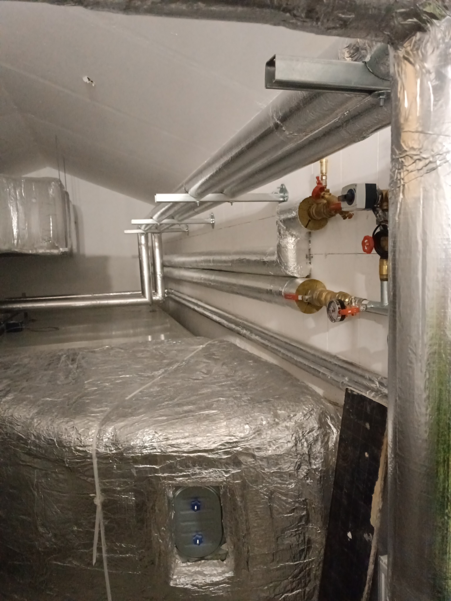 Izolowane rury instalacji grzewczej w piwnicy z zaworami i manometrami, widoczny zbiornik akumulacyjny obłożony folią izolacyjną.