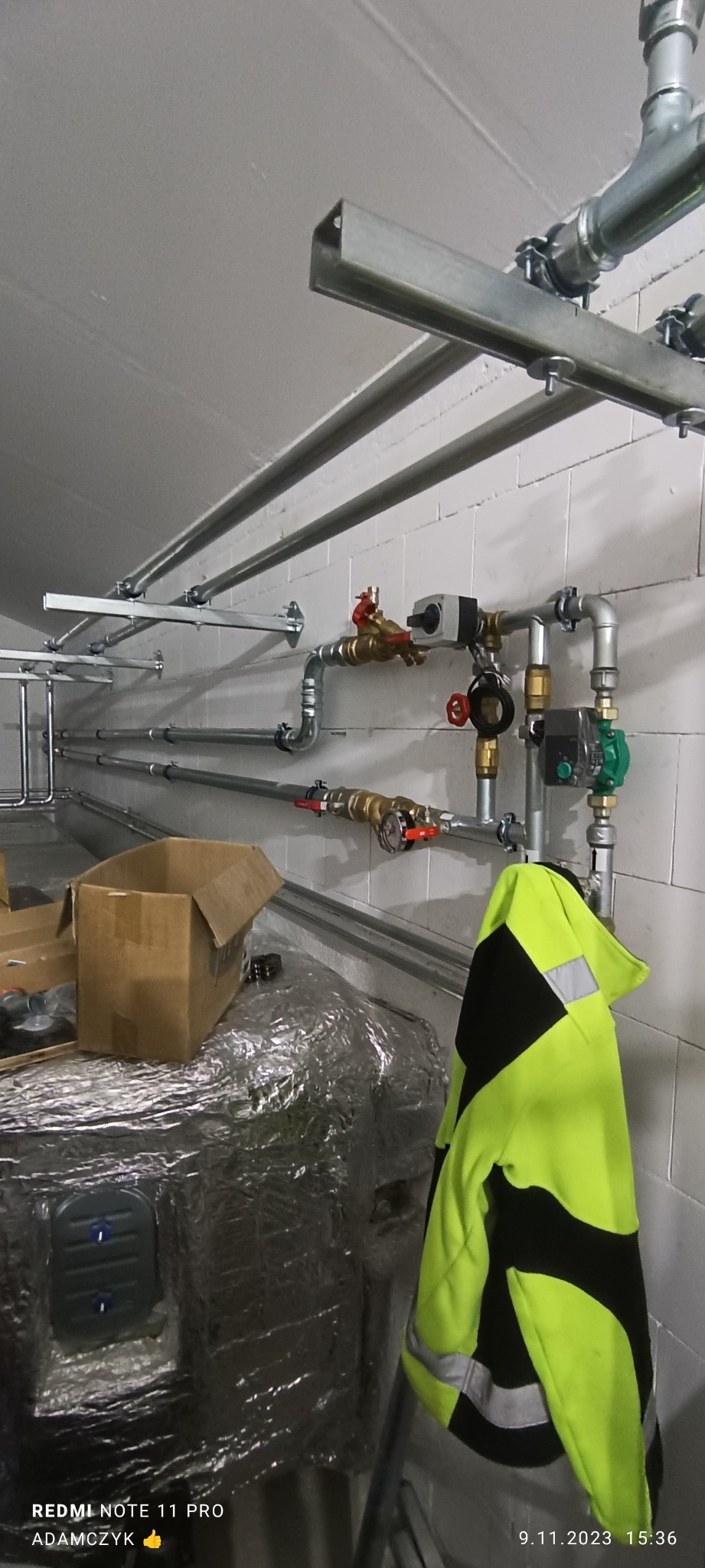 Szare rury instalacji grzewczej zamontowane na metalowych wspornikach przy białej, kafelkowanej ścianie. Widoczne zawory, pompy obiegowe i manometry. Na pierwszym planie, na drabinie, jaskrawożółta...