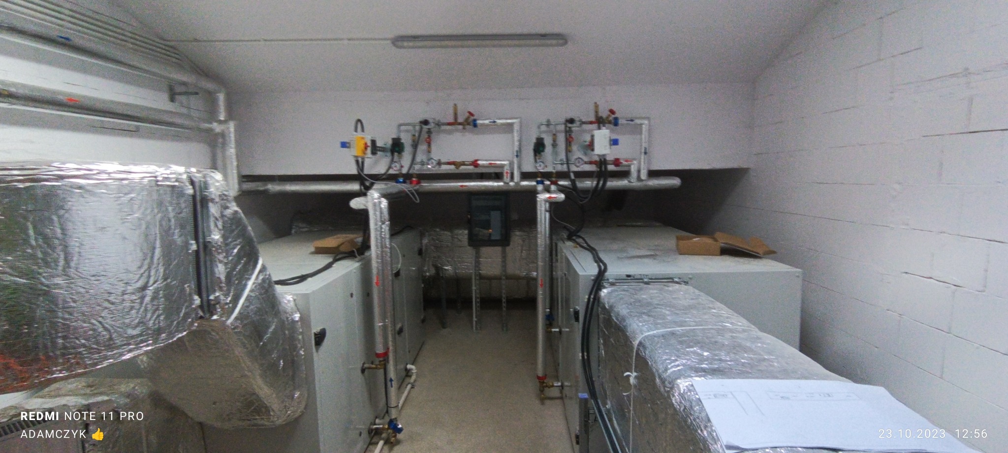 Dwa urządzenia wentylacyjne z rurami i zaworami w pomieszczeniu z białymi ścianami z cegieł, instalacja zabezpieczona folią aluminiową.