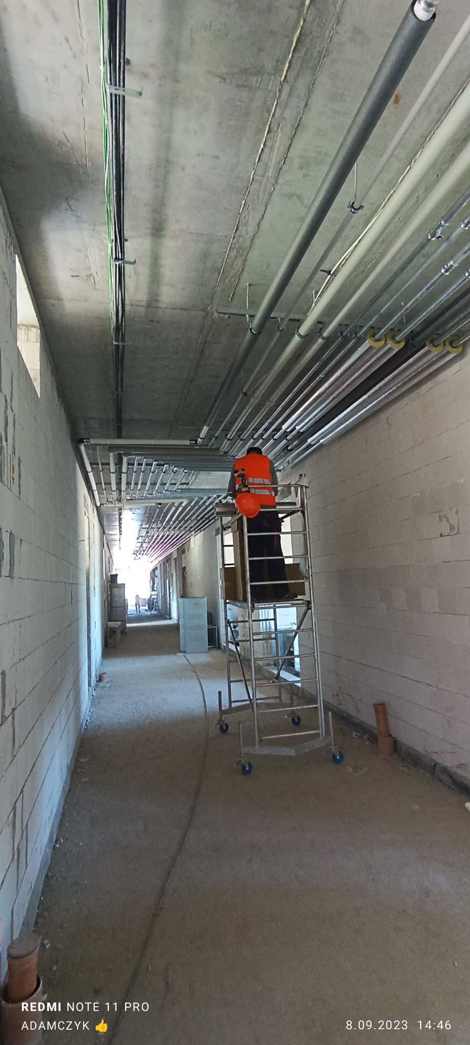 Instalator w pomarańczowej kamizelce na rusztowaniu montuje rury grzewcze pod sufitem w surowym wnętrzu budynku w trakcie budowy.