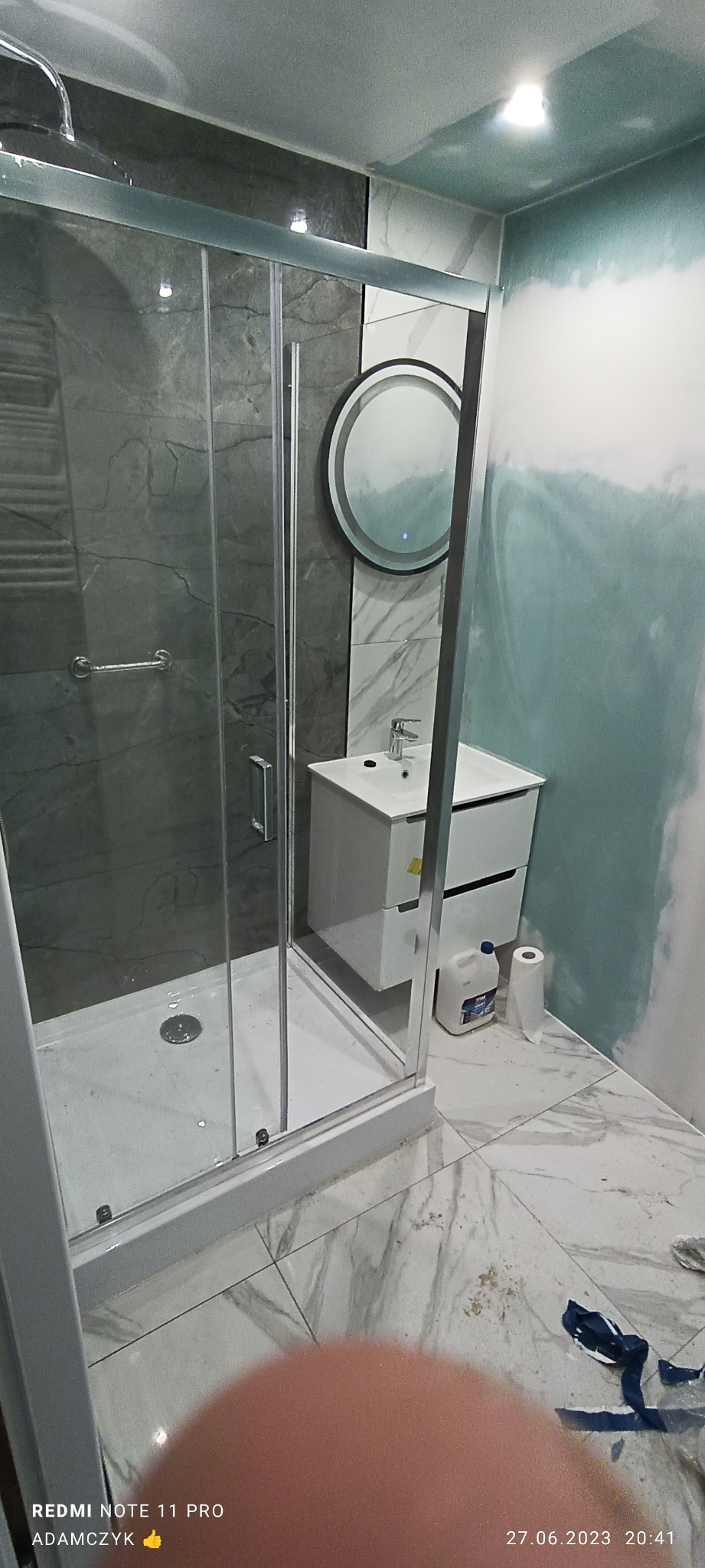 Nowoczesna łazienka w trakcie wykańczania: kabina prysznicowa ze szklanymi drzwiami, biała szafka umywalkowa z okrągłym lustrem z podświetleniem, marmurowe płytki podłogowe, ściana częściowo...