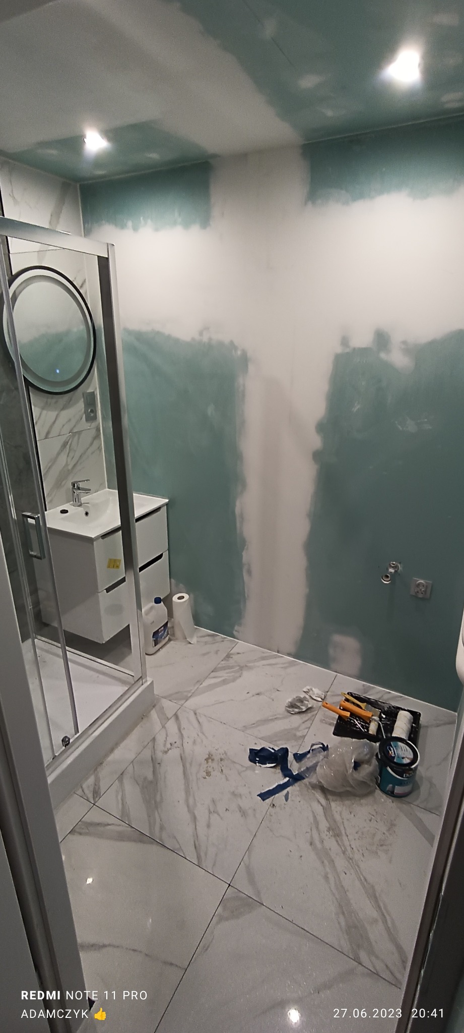 Łazienka w trakcie remontu: widoczne świeżo położone gładzie na ścianach, biała umywalka z szafką, kabina prysznicowa, oraz narzędzia malarskie i wiadro z farbą na marmurowej podłodze.