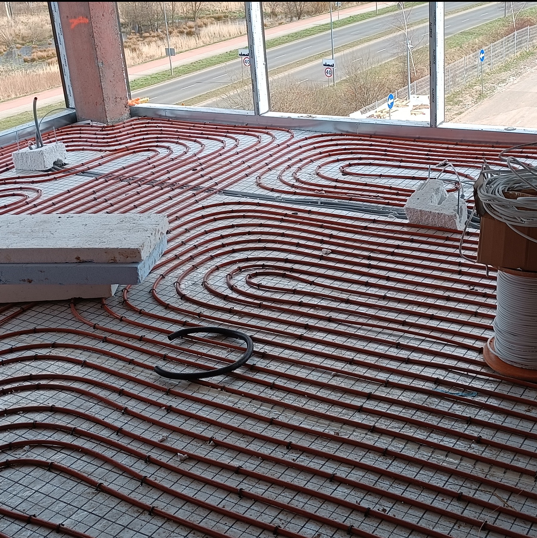 Montaż ogrzewania podłogowego w Bolkowie: czerwone rurki układane na siatce, widok z okna na ulicę zza betonowej kolumny.