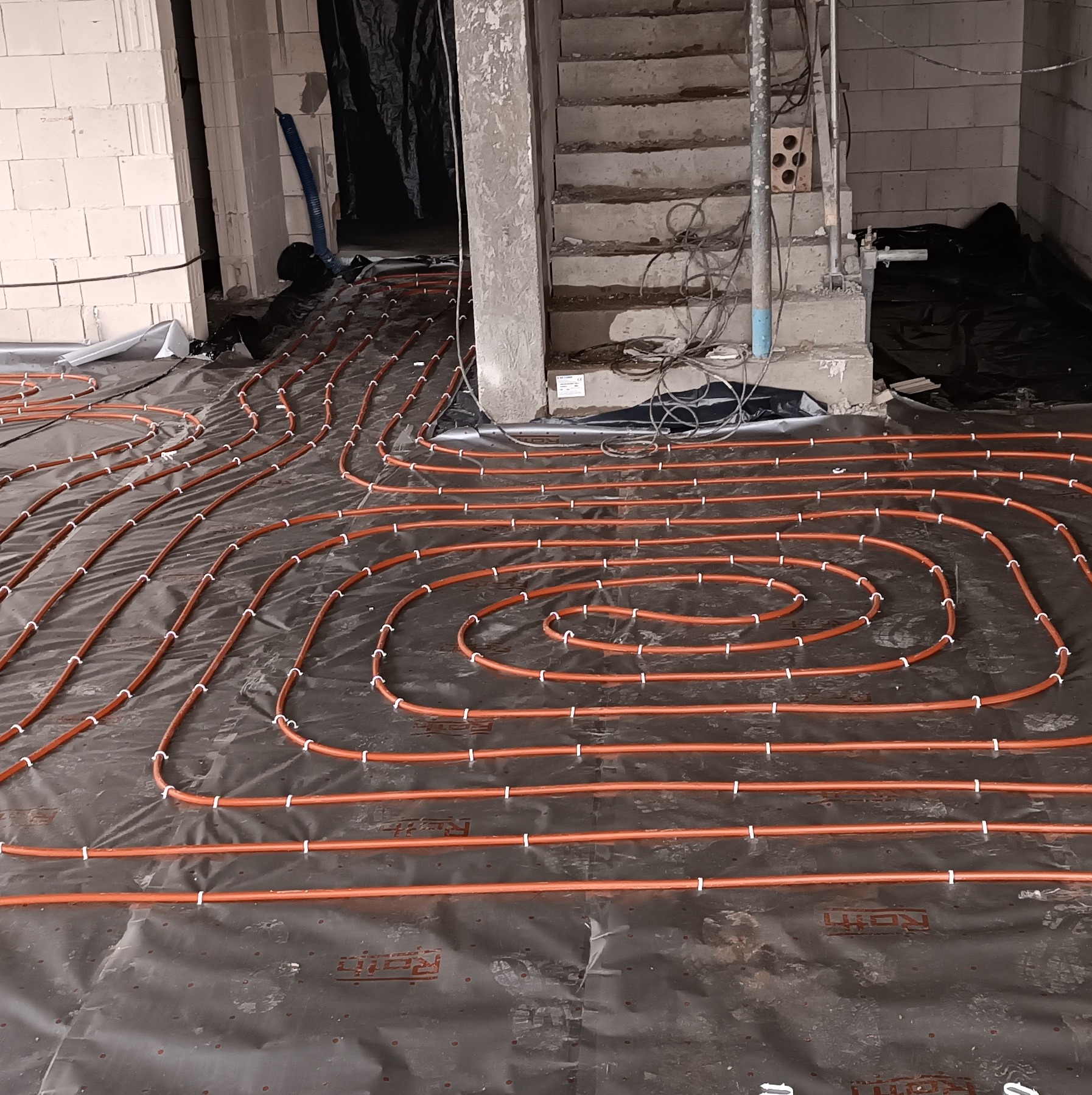 Pomarańczowe rury ogrzewania podłogowego ułożone spiralnie na czarnej folii izolacyjnej, białe zaciski mocujące, surowy stan wykończenia wnętrza, widoczne schody betonowe i ściany z pustaków.