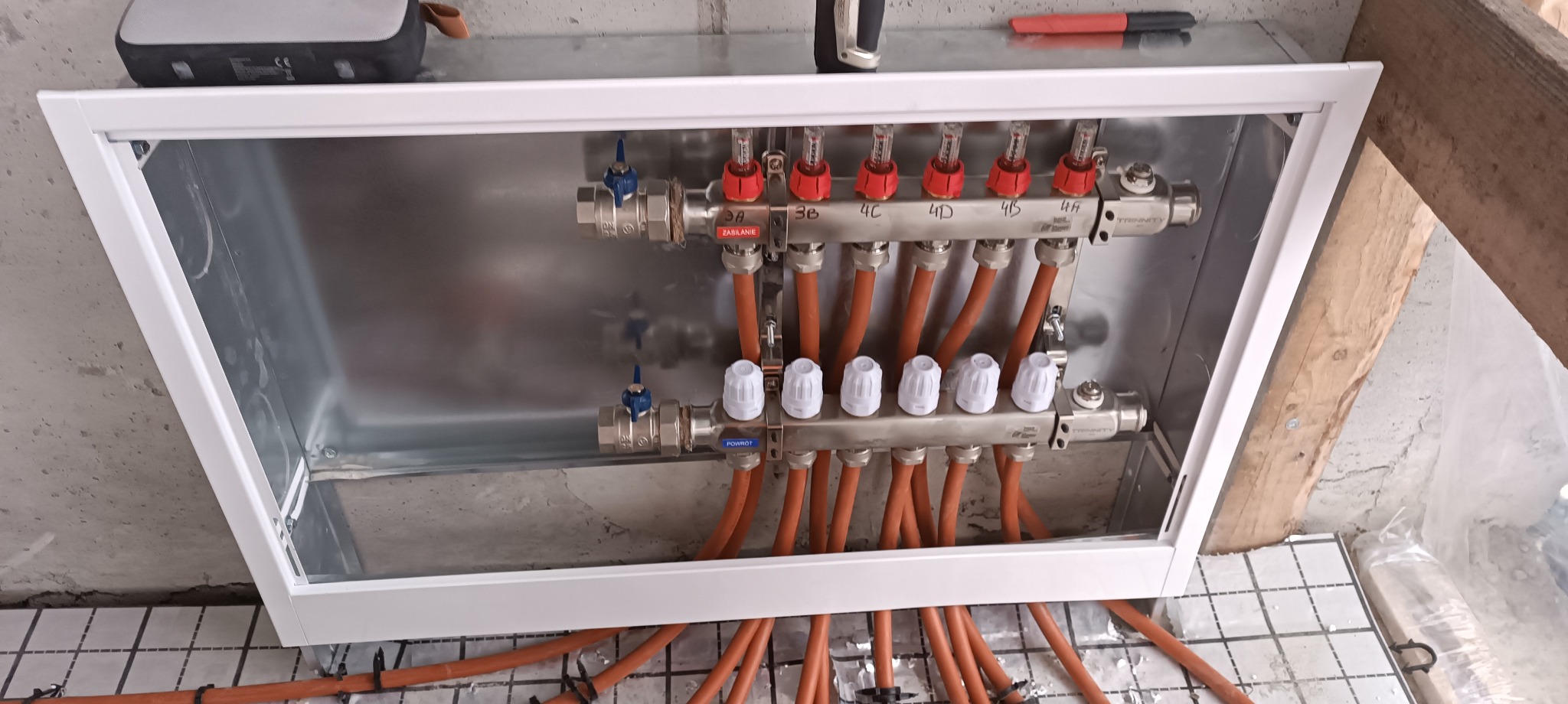 Rozdzielacz ogrzewania podłogowego w skrzynce instalacyjnej, widoczne rury w kolorze pomarańczowym, zawory i liczniki przepływu, zamontowany na podłodze z izolacją.