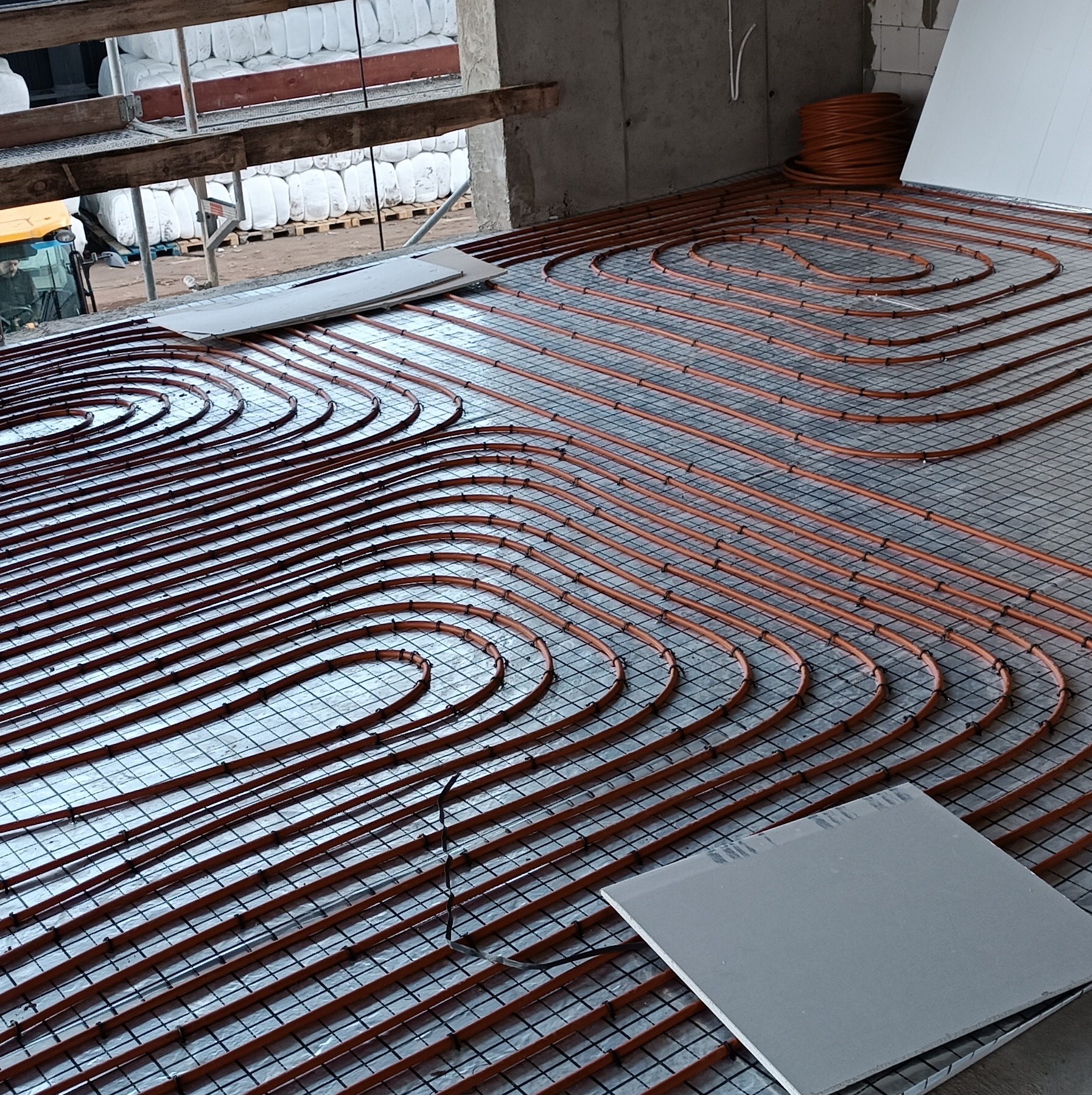 Montaż ogrzewania podłogowego: pomarańczowe rury rozłożone na metalowej siatce w surowym wnętrzu budynku w trakcie budowy, widoczne płyty izolacyjne i zwój rur.