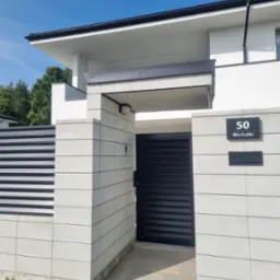 Nowoczesna brama wjazdowa z paneli stalowych w kolorze czarnym, umieszczona w jasnoszarym ogrodzeniu z bloczków betonowych, z numerem domu '50 Michałki' i skrzynką na listy.