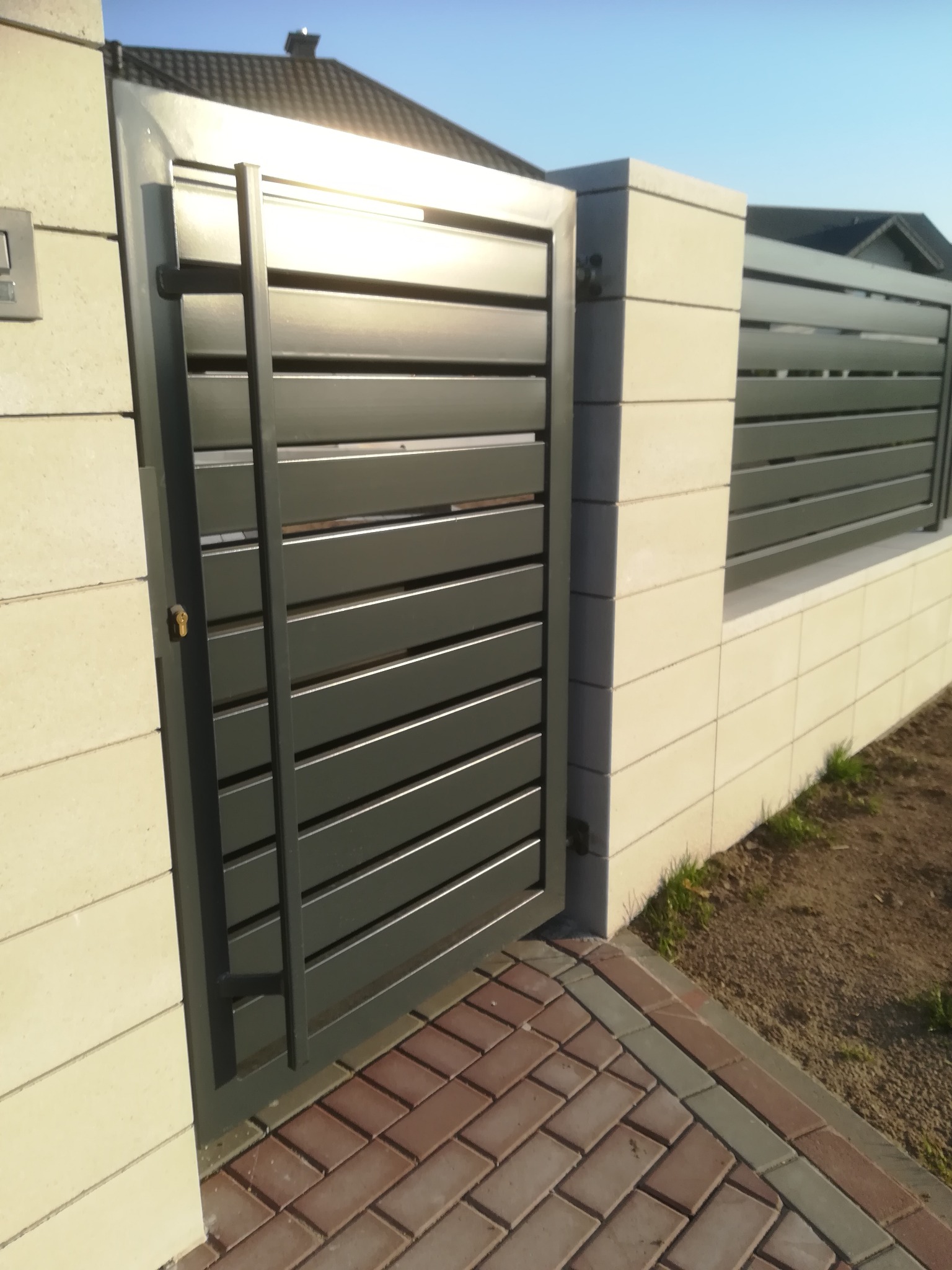 Nowoczesna brama wjazdowa z poziomych profili w kolorze grafitowym, osadzona w słupkach z bloczków betonowych, z widoczną klamką i fragmentem ogrodzenia panelowego w tle.