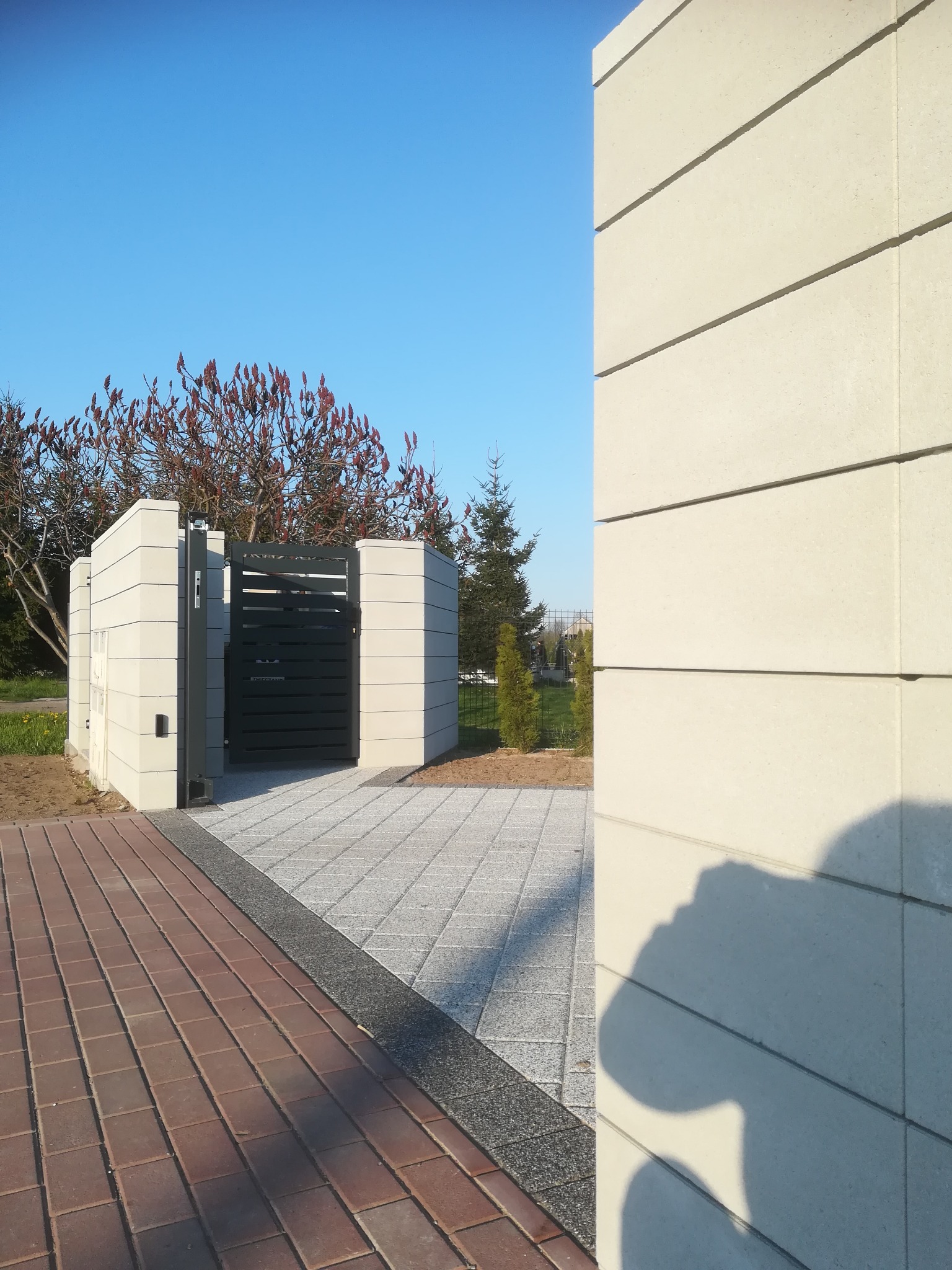 Nowoczesna, pozioma brama wjazdowa w kolorze grafitowym, osadzona w jasnych, betonowych słupkach, z widocznym fragmentem ceglanej ścieżki i betonowej nawierzchni, na tle błękitnego nieba i zieleni...