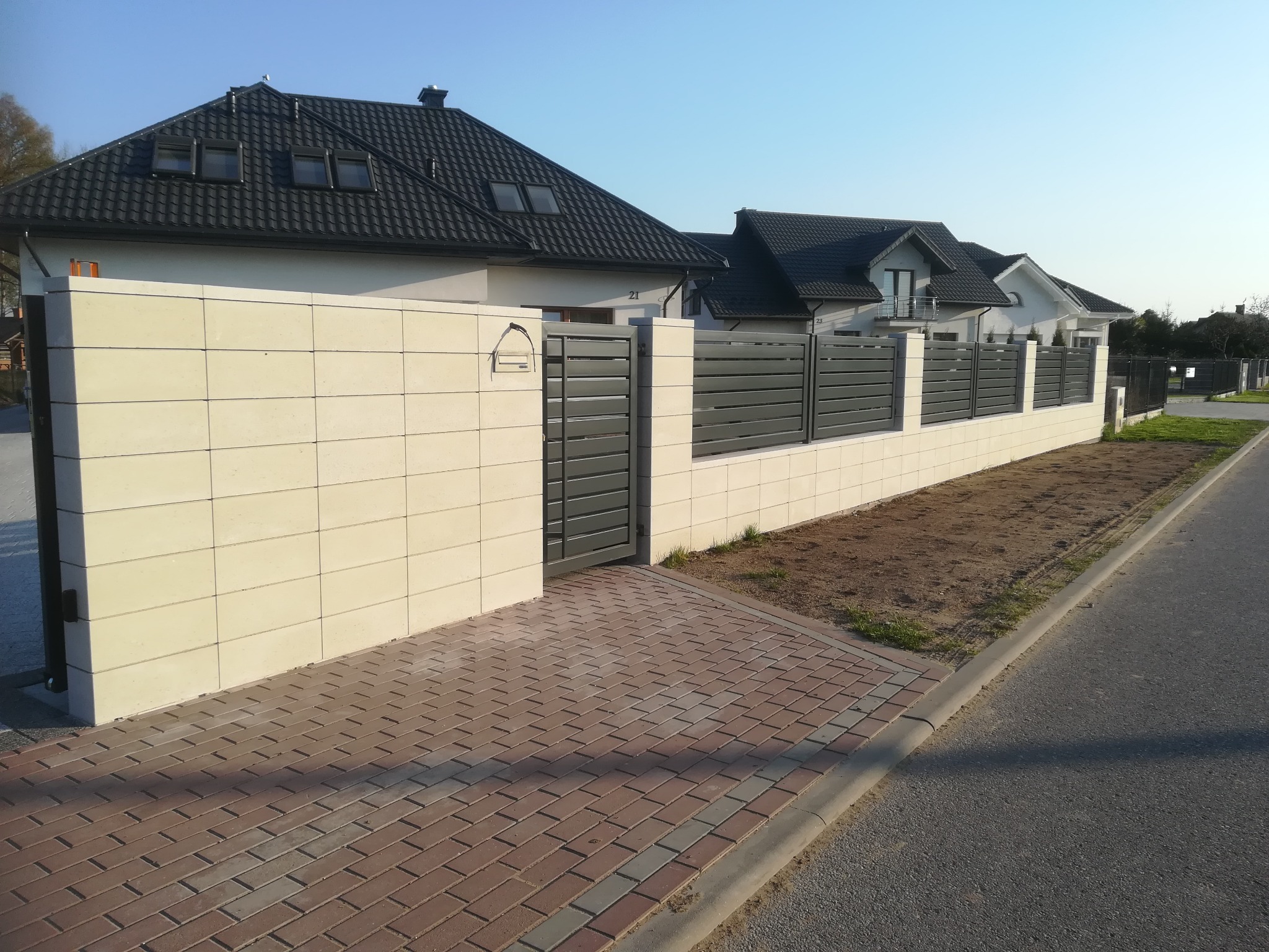 Nowoczesne ogrodzenie posesji z przesuwną bramą, wykonane z paneli poziomych w kolorze grafitowym, osadzone na jasnych, modułowych słupkach betonowych, z widocznym fragmentem chodnika z kostki...