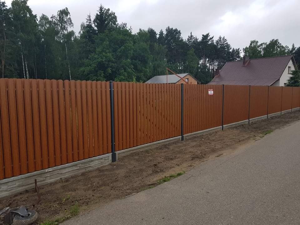 Długie ogrodzenie panelowe z brązowych pionowych desek i szarych słupków, zamontowane na betonowej podmurówce, widoczne przy drodze asfaltowej na tle drzew i domu.