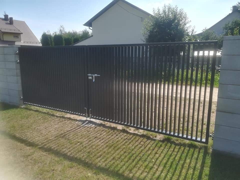Nowoczesna, grafitowa brama wjazdowa z pionowych profili, zamontowana między betonowymi słupkami na tle domu jednorodzinnego i trawnika.