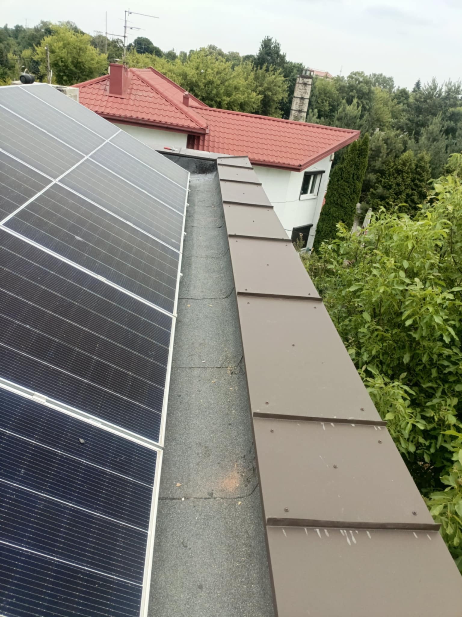 Widok z góry na fragment dachu z panelami słonecznymi i brązowymi obróbkami blacharskimi, w tle dom z czerwoną dachówką i zielona roślinność.