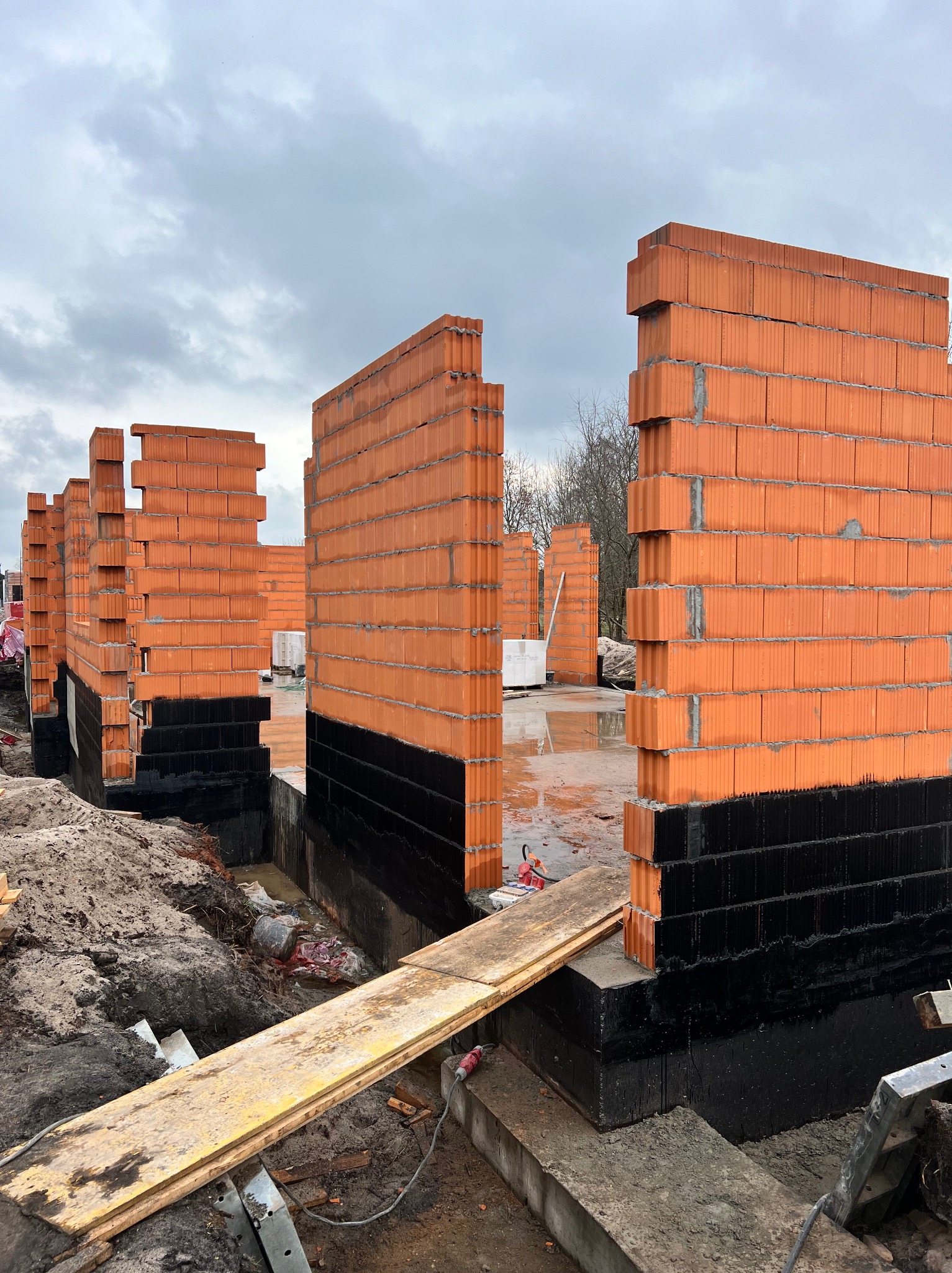 Wznoszenie ścian parteru domu z czerwonej ceramiki poryzowanej, zabezpieczonych czarną hydroizolacją fundamentów, widok na plac budowy w pochmurny dzień.