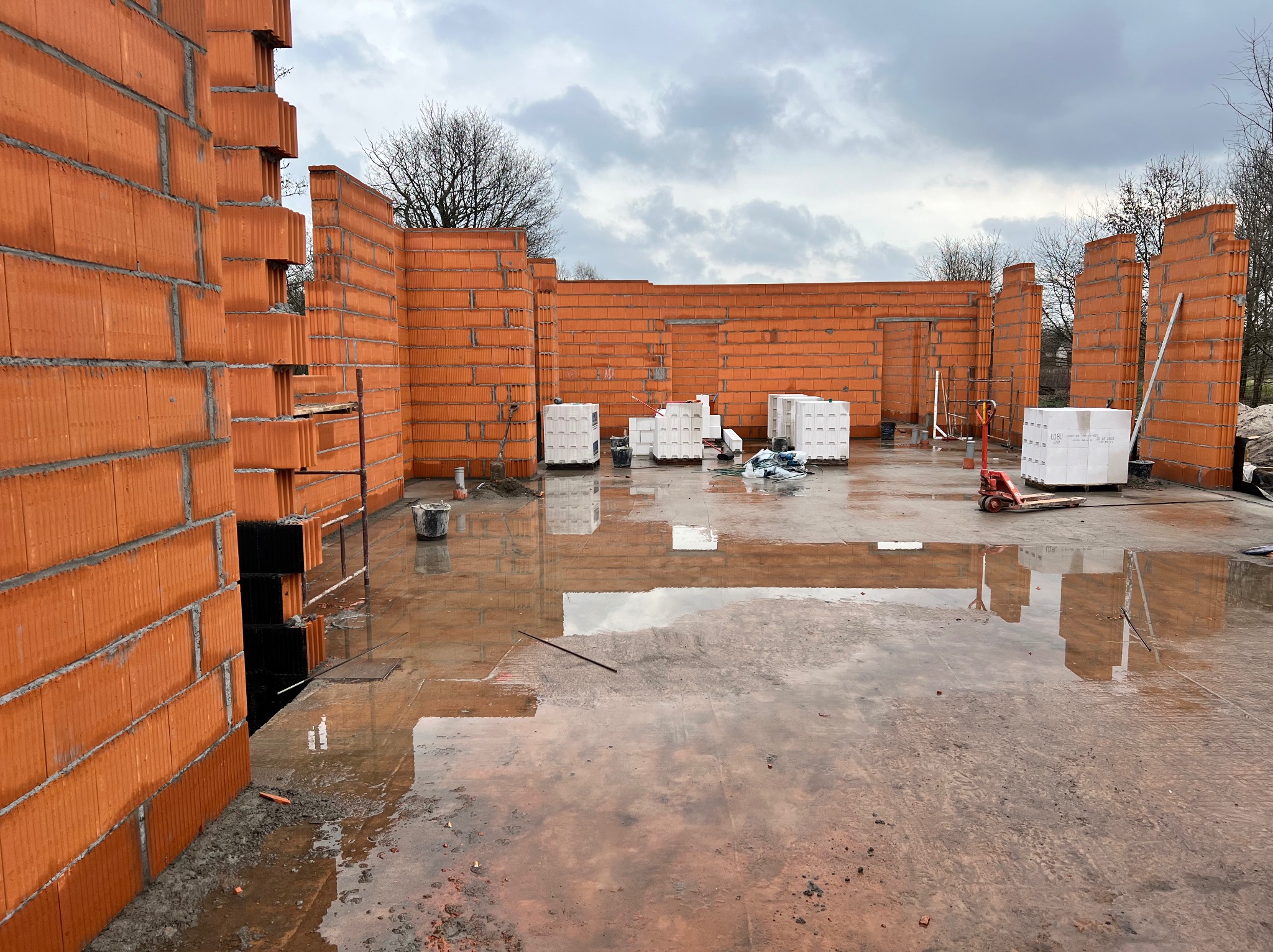 Wnętrze domu w trakcie budowy z czerwonej cegły ceramicznej, widoczne ściany, palety cegieł, mokra posadzka i pochmurne niebo w tle.