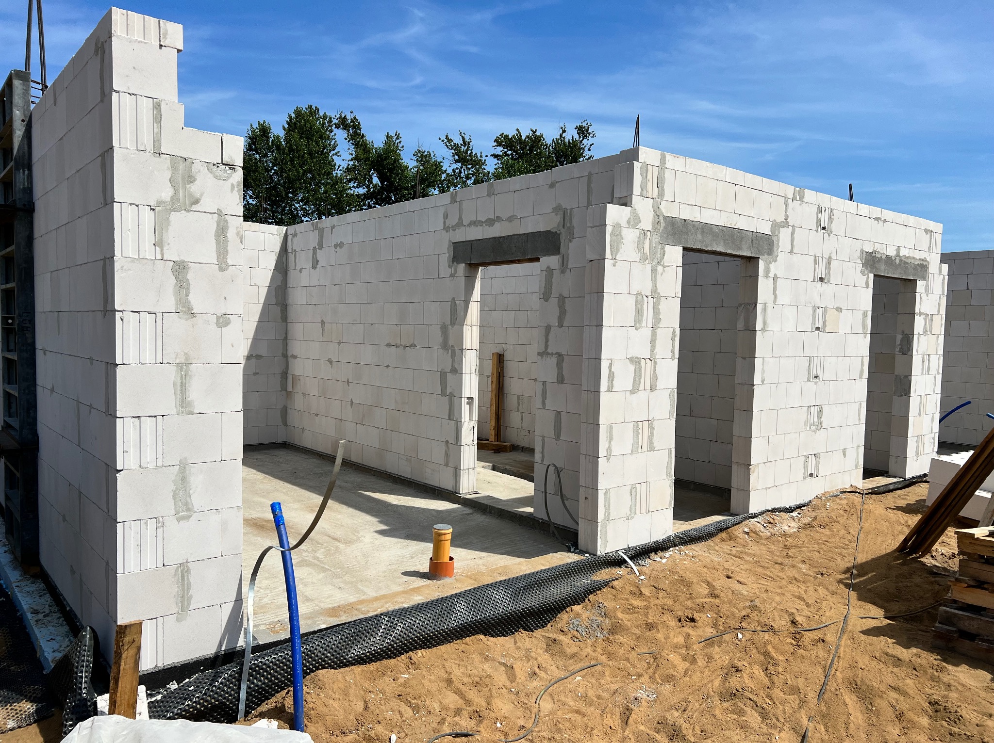Surowy stan domu z białych bloczków, widoczne otwory okienne i drzwiowe, grunt wokół budynku częściowo pokryty czarną folią drenażową, niebo błękitne w tle.