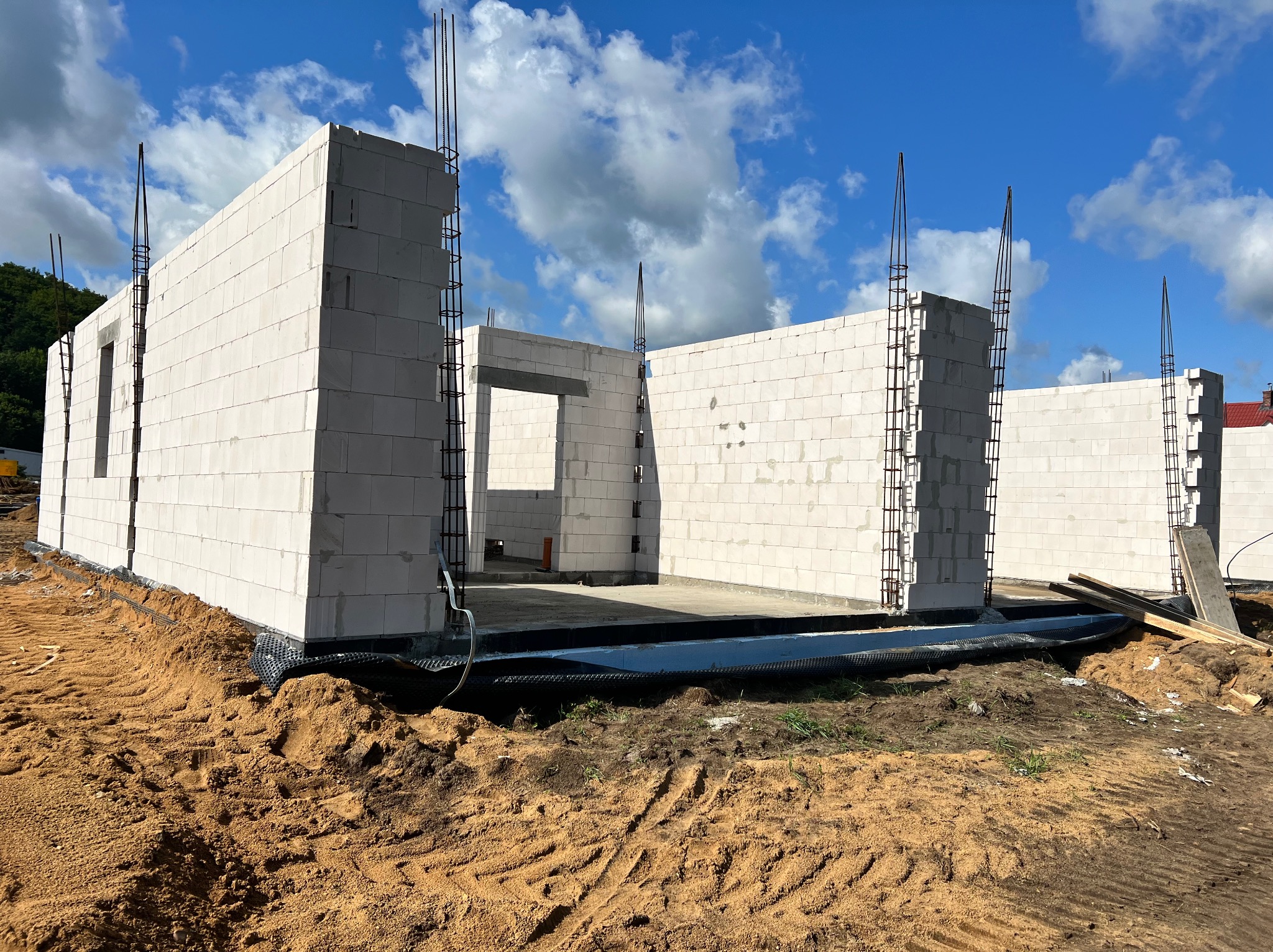 Wznoszone ściany domu z białych bloczków, widoczne zbrojenia w narożnikach, teren wokół w stanie surowym, ślady opon na piasku.