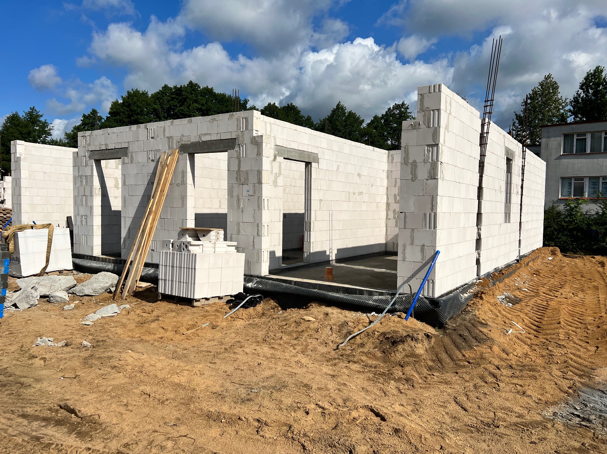Wznoszone ściany domu z białych bloczków z betonu komórkowego, widoczne zbrojenia pionowe, teren wokół w stanie surowym.