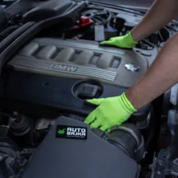 Silnik BMW podczas prac serwisowych, dłonie w jaskrawozielonych rękawicach umieszczone na pokrywie silnika, widoczna naklejka z logo firmy na obudowie filtra powietrza.