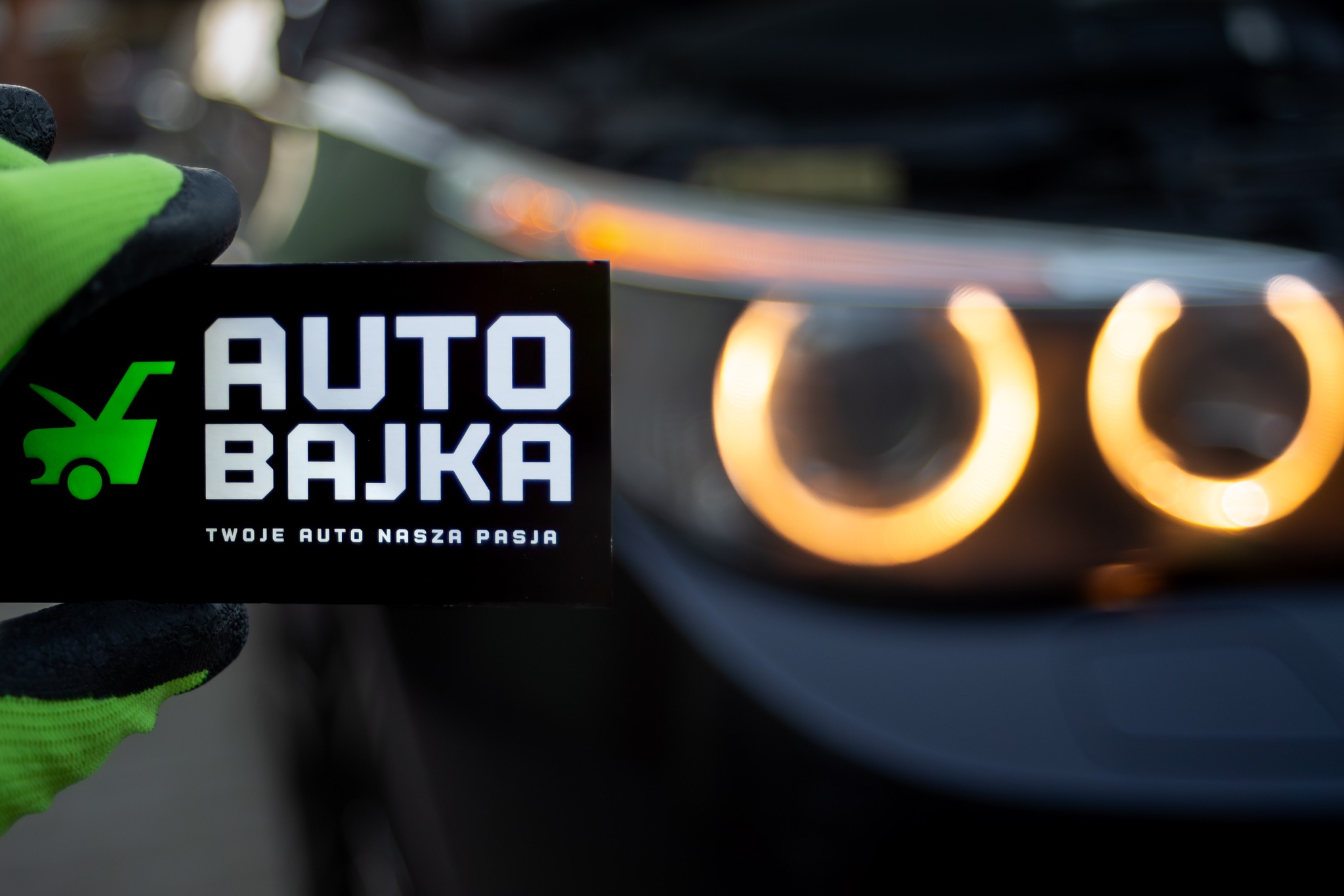Karta z logo 'Auto Bajka' trzymana w rękawicy roboczej na tle rozświetlonych reflektorów samochodowych.
