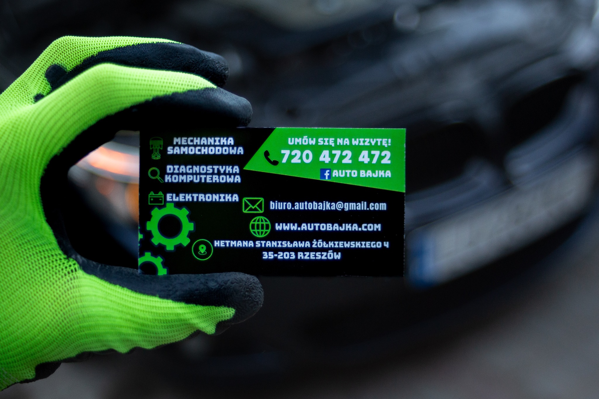 Zielona rękawica ochronna trzyma czarny wizytownik z zielonymi napisami oferujący usługi: mechanika samochodowa, diagnostyka komputerowa i elektronika. W tle fragment rozmytego pojazdu.