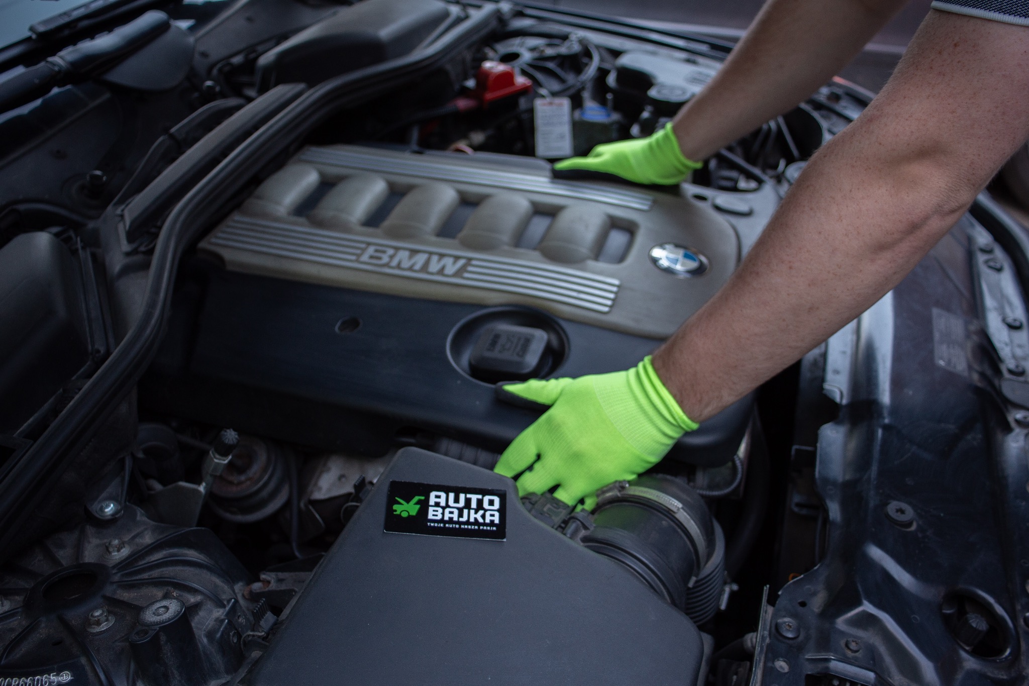 Silnik BMW podczas prac serwisowych, dłonie w jaskrawozielonych rękawicach umieszczone na pokrywie silnika, widoczna naklejka z logo firmy na obudowie filtra powietrza.