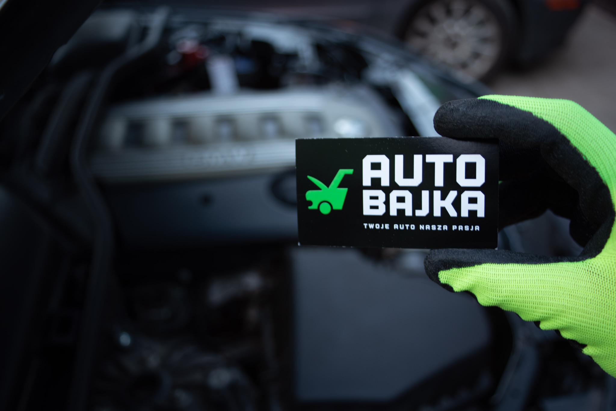Silnik samochodowy widziany przez otwartą maskę, w tle zamazane koło, w pierwszym planie dłoń w neonowo-zielonej rękawicy trzymająca wizytówkę firmy 'Auto Bajka'.