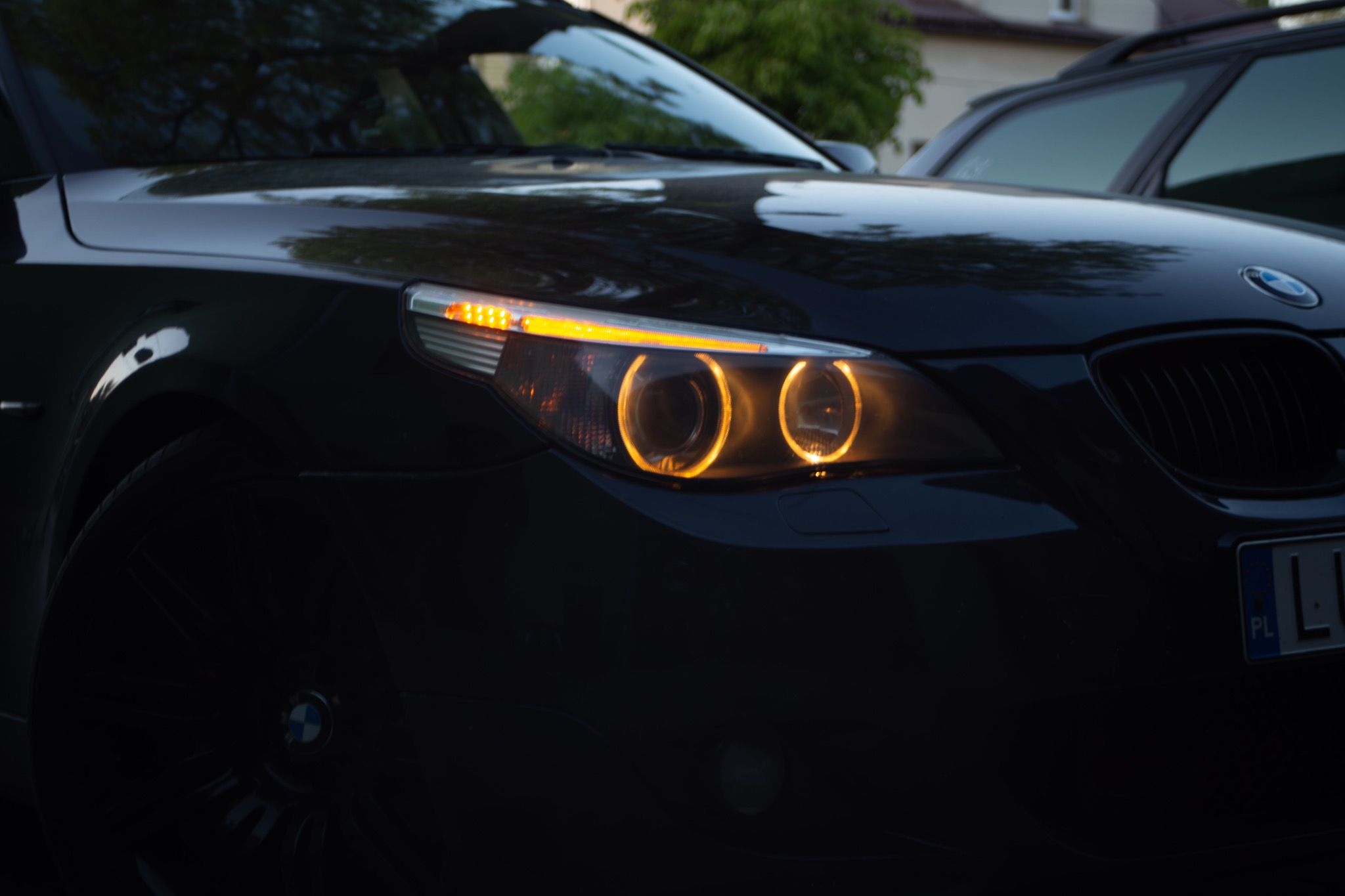 Zbliżenie na przedni reflektor BMW z charakterystycznymi okrągłymi światłami pozycyjnymi, lekko przyciemniony obraz z widocznym fragmentem tablicy rejestracyjnej i zarysem innego pojazdu w tle.