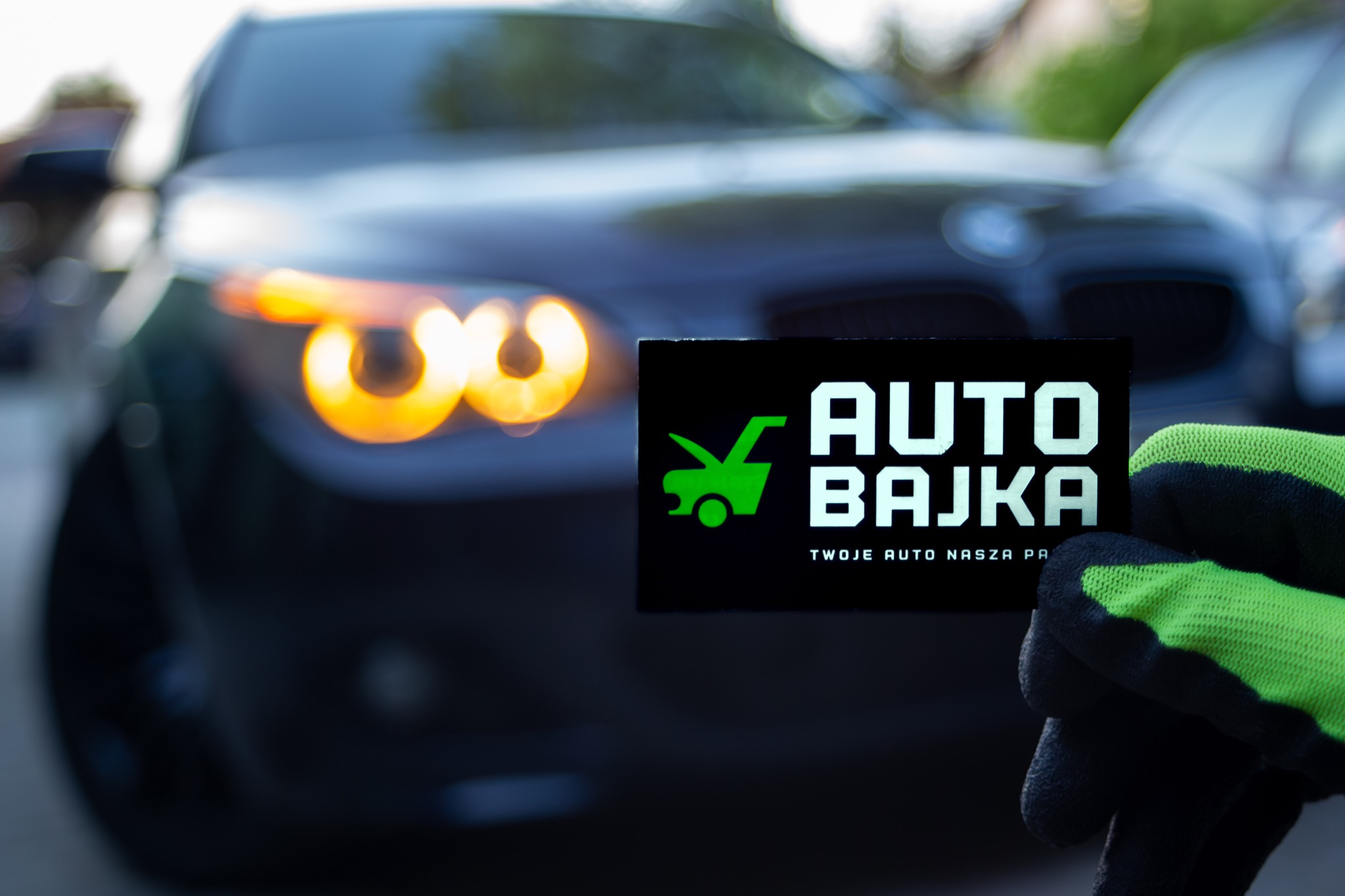 Ciemny samochód z włączonymi światłami w tle, na pierwszym planie dłoń w roboczej rękawicy trzymająca czarną tabliczkę z zielonym logo 'Auto Bajka' i tekstem 'Twoje auto nasza pasja'.