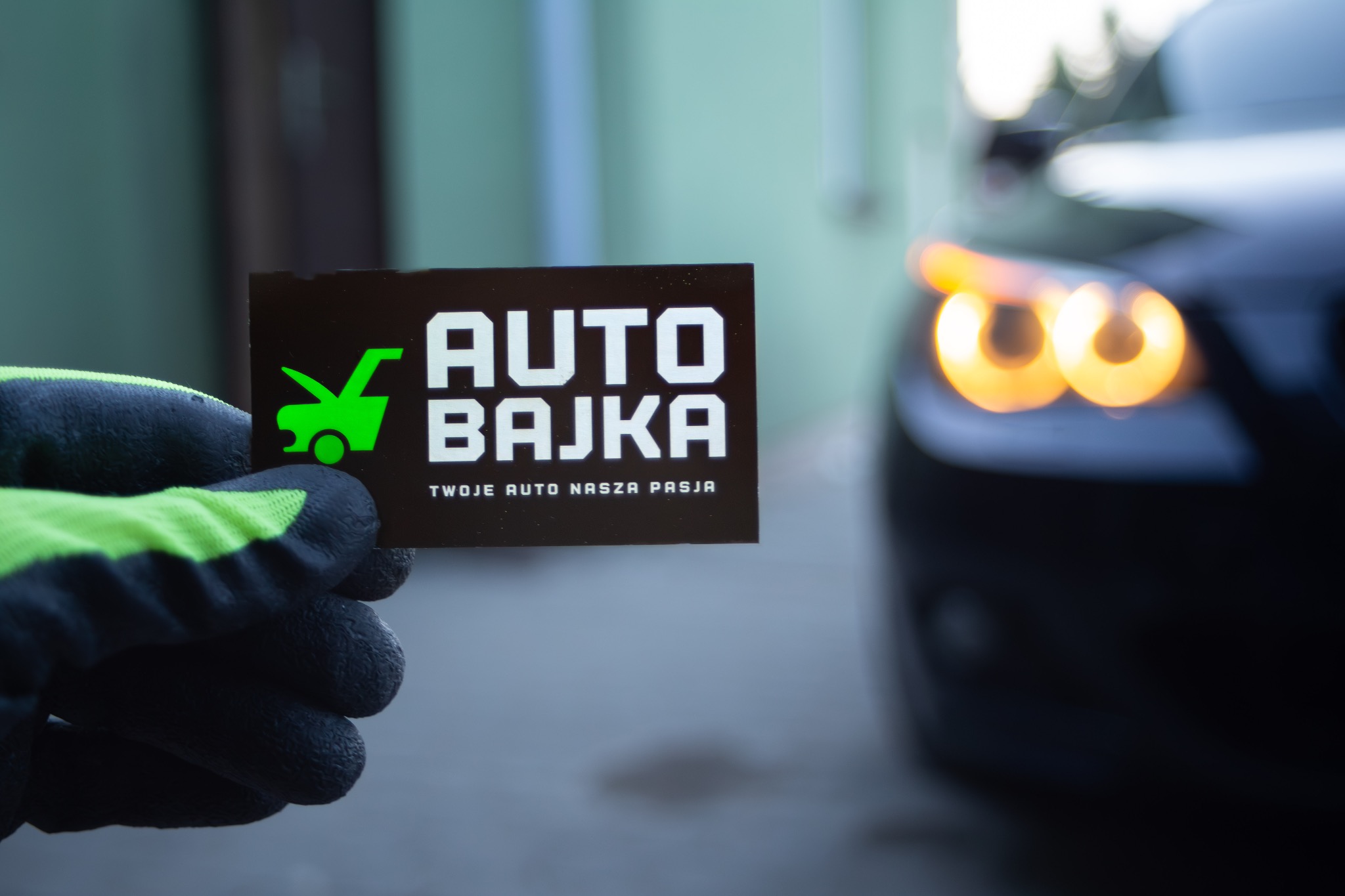 Czarna wizytówka z logo 'AUTO BAJKA' trzymana w dłoni w roboczej rękawicy z zielonymi elementami; w tle rozmyte światła samochodu.
