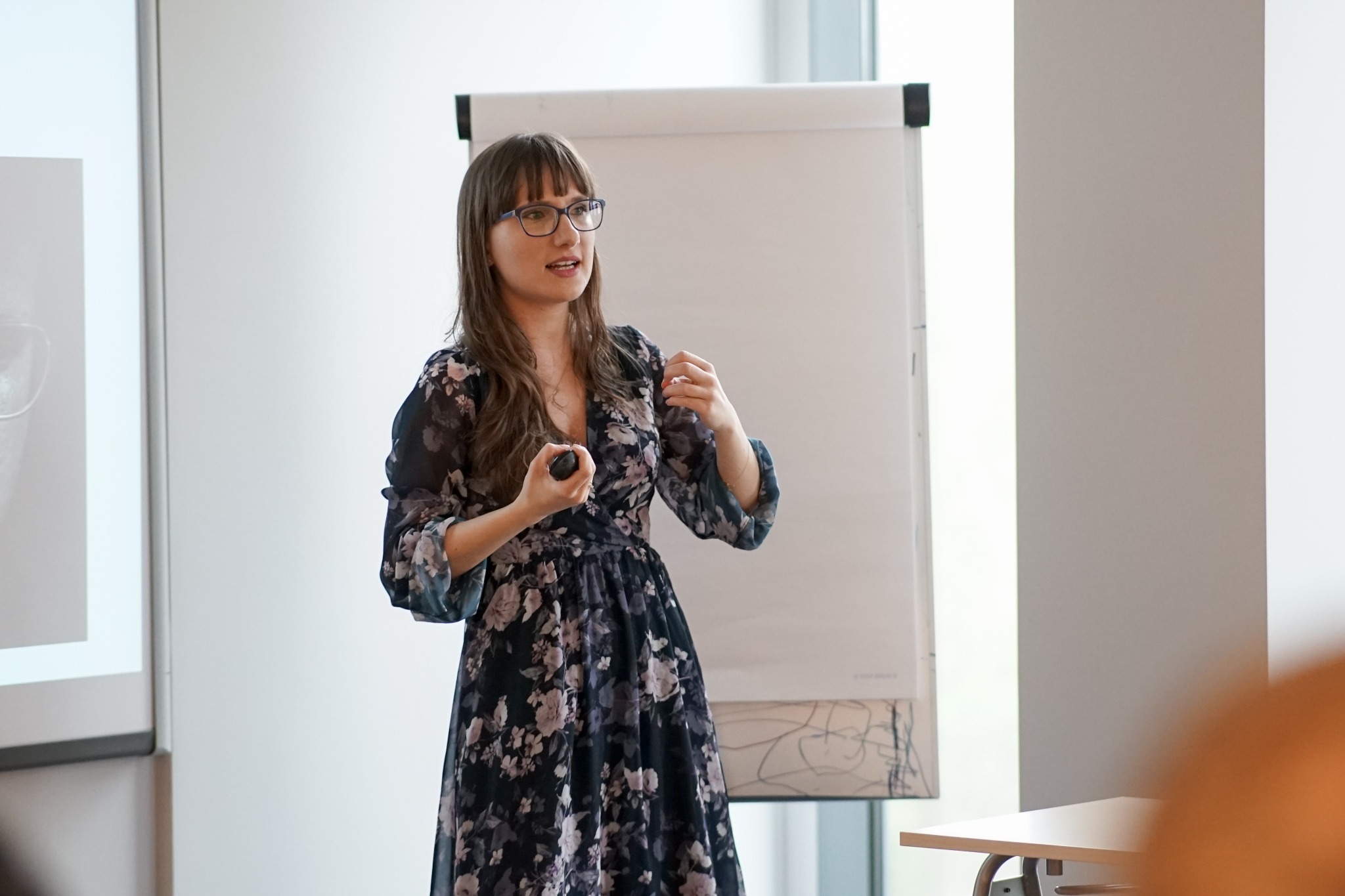 Kobieta w okularach i kwiecistej sukience prezentuje coś trzymając pilot do prezentacji, przed nią biała tablica flipchart i ekran z częściowo widocznym zdjęciem.