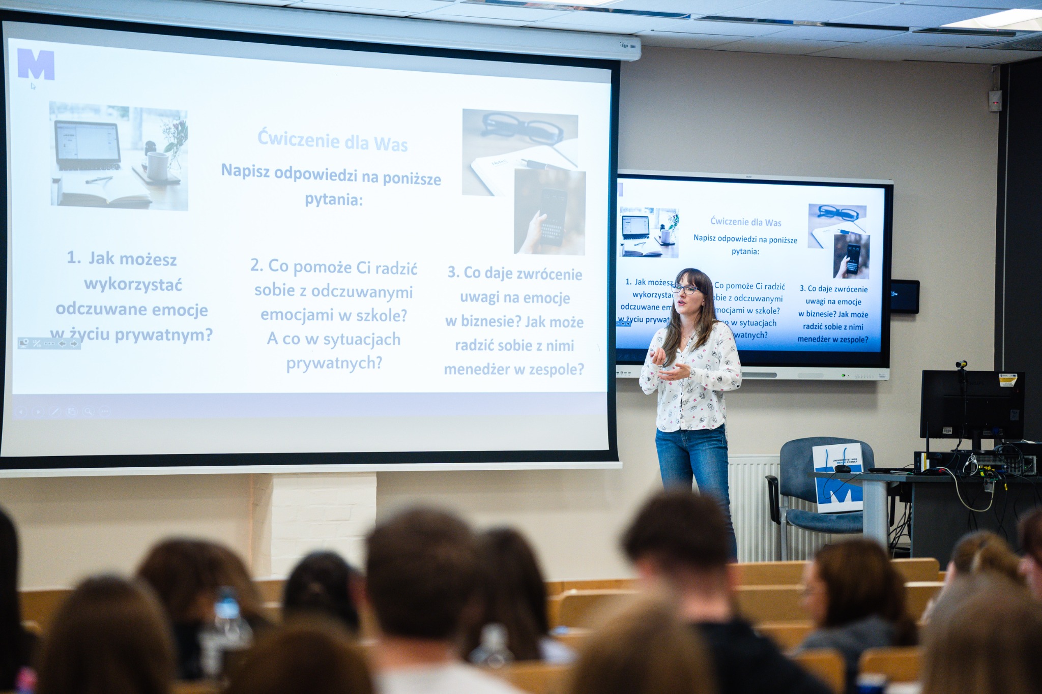 Kobieta w okularach prezentuje na sali wykładowej slajd z pytaniami dotyczącymi radzenia sobie z emocjami w życiu prywatnym i zawodowym, widok zza pleców słuchaczy.