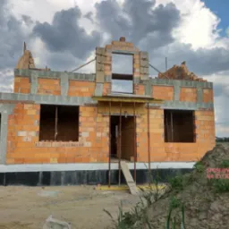 Wznoszenie ścian domu jednorodzinnego z cegły, widoczne otwory okienne i drzwiowe, szare niebo w tle, etap budowy w stanie surowym otwartym.