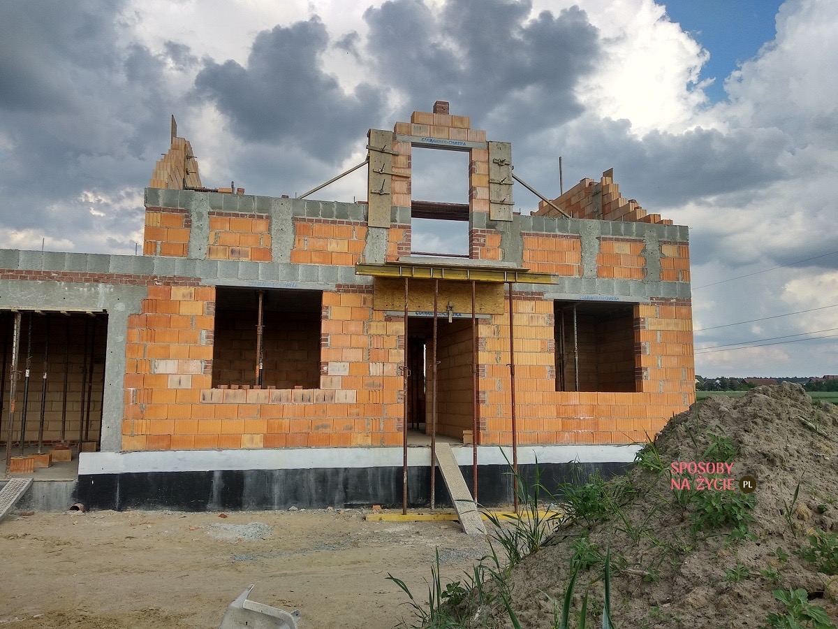 Wznoszenie ścian domu jednorodzinnego z cegły, widoczne otwory okienne i drzwiowe, szare niebo w tle, etap budowy w stanie surowym otwartym.