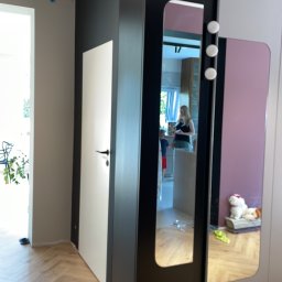 YA COMPANY SPÓŁKA Z OGRANICZONĄ ODPOWIEDZIALNOŚCIĄ - Fragment wykończonego wnętrza mieszkania: parkiet w jodełkę, ściana pomalowana na czarno, białe drzwi, szafa z lustrem, w którym odbija się wnętrze kuchni.