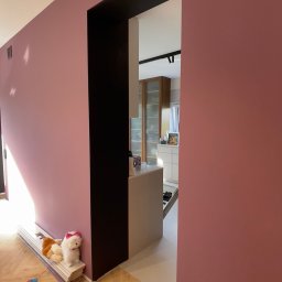 YA COMPANY SPÓŁKA Z OGRANICZONĄ ODPOWIEDZIALNOŚCIĄ - Ściana pomalowana na kolor brudny róż z czarną wnęką drzwiową, widok na wnętrze kuchni z drewnianymi szafkami i białą wyspą, na podłodze parkiet w jodełkę, przy ścianie leżą pluszowe zabawki.