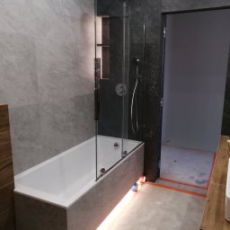 YA COMPANY SPÓŁKA Z OGRANICZONĄ ODPOWIEDZIALNOŚCIĄ - Nowoczesna łazienka z wanną obudowaną płytkami imitującymi beton, szklanym parawanem nawannowym i podświetleniem LED u dołu. Widoczna również deszczownica i fragment umywalki z blatem z drewna.
