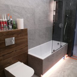 YA COMPANY SPÓŁKA Z OGRANICZONĄ ODPOWIEDZIALNOŚCIĄ - Nowoczesna łazienka z wanną obudowaną szarą płytką, szklanym parawanem nawannowym, podświetleniem LED i wiszącą toaletą z drewnopodobną zabudową ścienną. Widoczne silikony i akcesoria na półce.