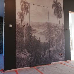 YA COMPANY SPÓŁKA Z OGRANICZONĄ ODPOWIEDZIALNOŚCIĄ - Pokój w trakcie remontu z ciemną ścianą, na której umieszczono fototapetę z motywem tropikalnego krajobrazu i ukrytymi drzwiami. Podłoga zabezpieczona folią i taśmą malarską.