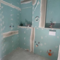 YA COMPANY SPÓŁKA Z OGRANICZONĄ ODPOWIEDZIALNOŚCIĄ - Niedokończona łazienka z płytami gipsowo-kartonowymi, widoczne instalacje wodne i elektryczne, przygotowana do dalszych prac wykończeniowych, z pozostawionymi narzędziami w tle.