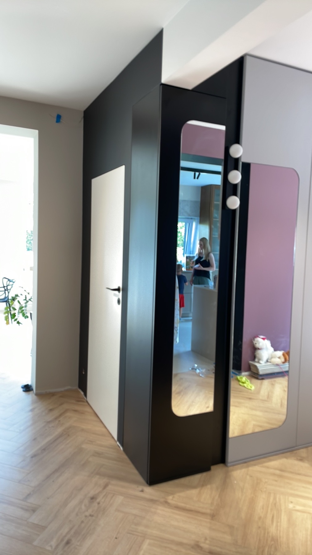 Fragment wykończonego wnętrza mieszkania: parkiet w jodełkę, ściana pomalowana na czarno, białe drzwi, szafa z lustrem, w którym odbija się wnętrze kuchni.