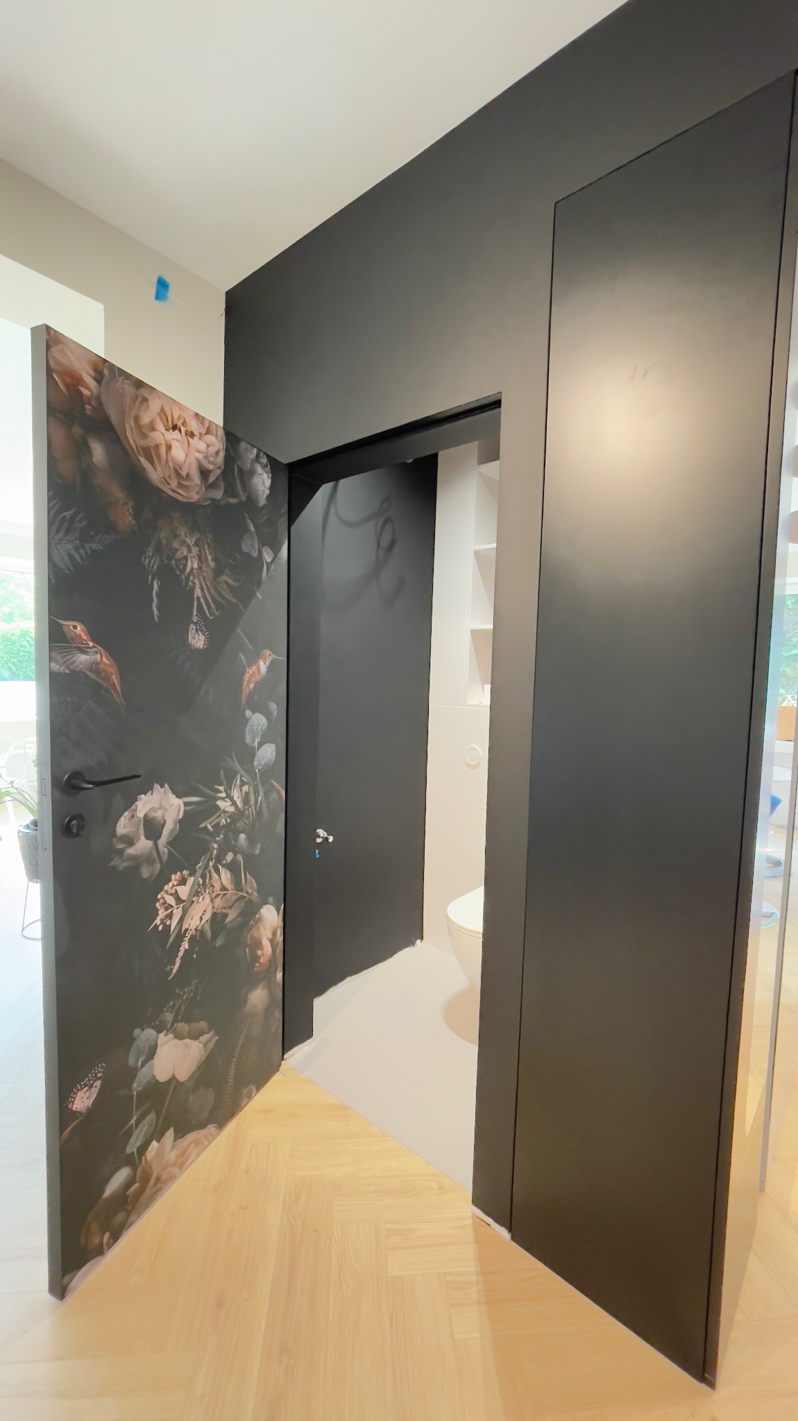 Otwarta, czarna szafa wnękowa z fototapetą o motywie kwiatowym i ptaków, przysłaniająca wejście do toalety w mieszkaniu w Warszawie.