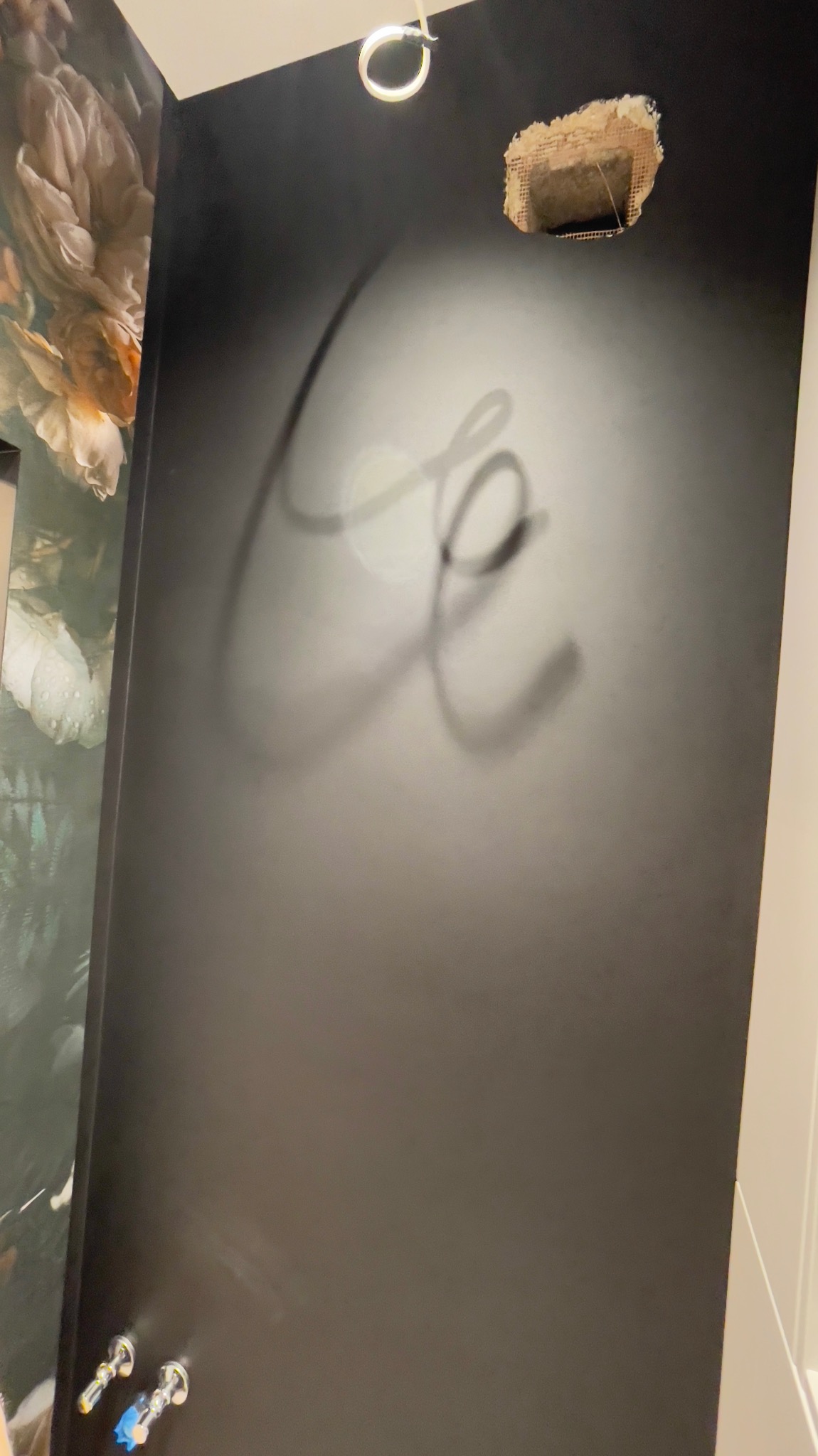 Ciemna ściana z niedokończoną instalacją elektryczną i zamalowanym napisem, fragment tapety w kwiaty po lewej stronie i instalacja wodna u dołu.