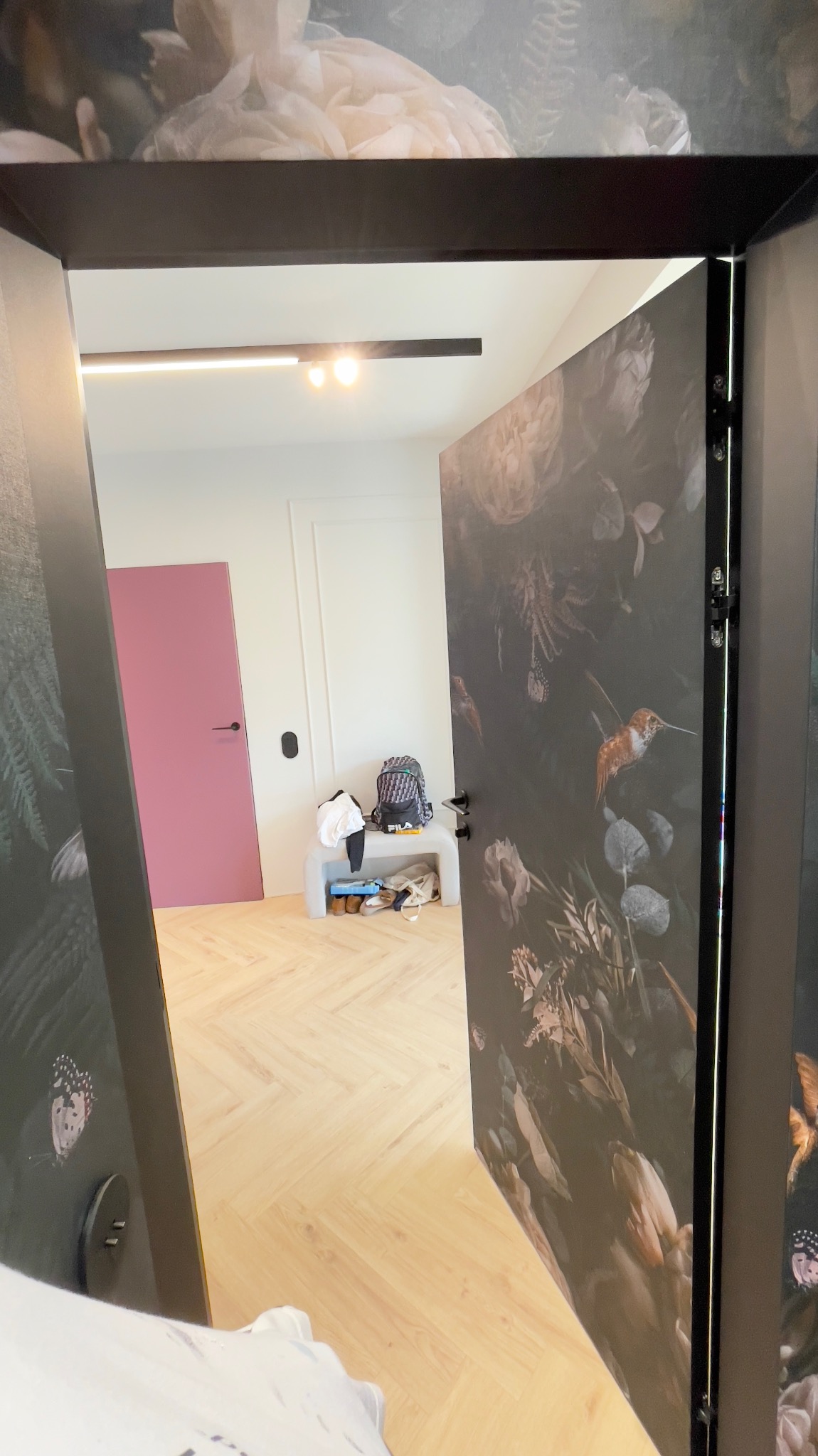 Widok na pokój przez otwarte drzwi z motywem kwiatowym, różowe drzwi w tle, podłoga w jodełkę, minimalistyczne oświetlenie sufitowe.