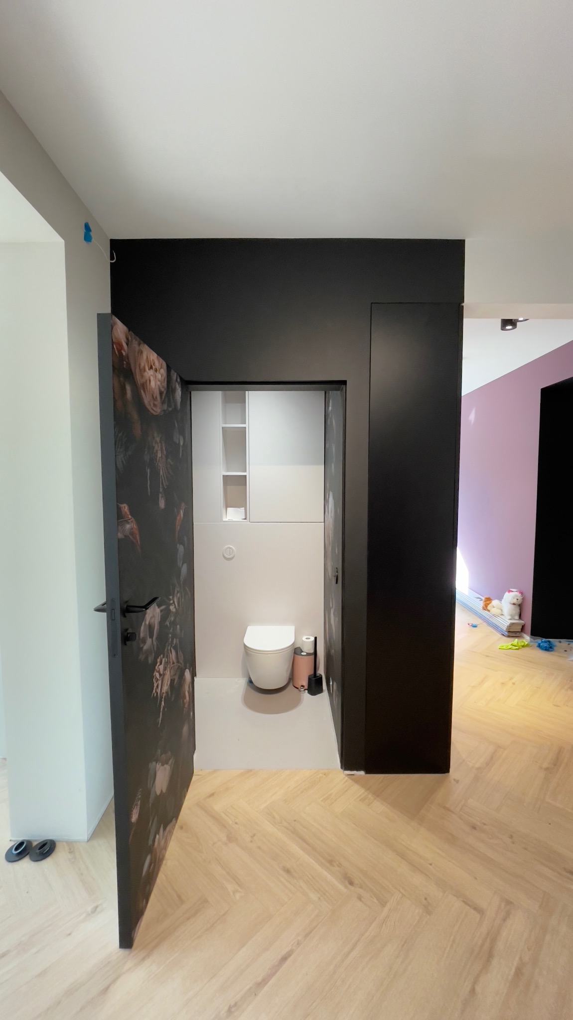 Nowoczesna toaleta z czarną zabudową, otwartymi drzwiami z motywem kwiatowym, jasnymi płytkami i drewnianą podłogą ułożoną w jodełkę.