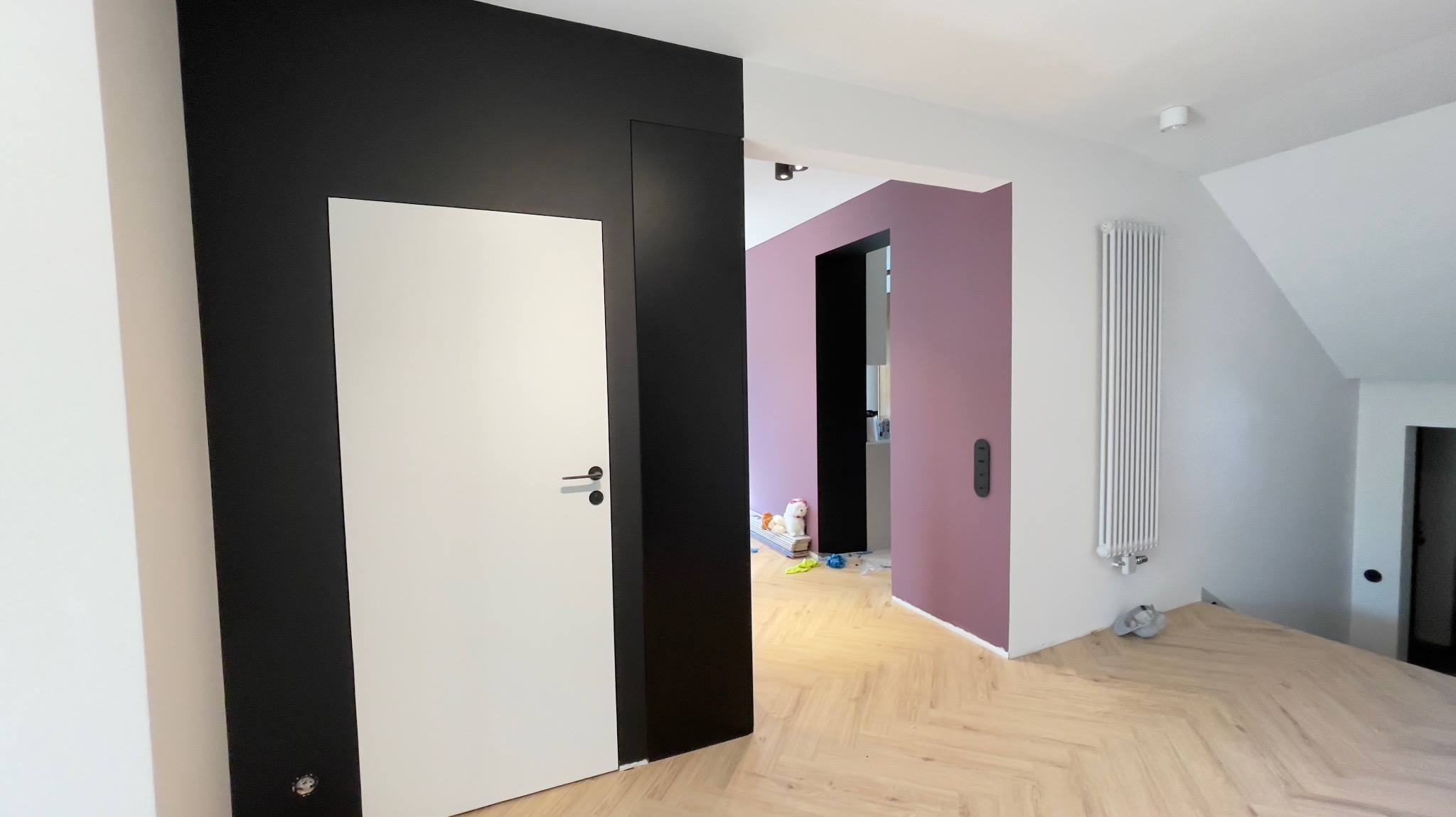Nowoczesne wnętrze z kontrastującymi kolorami ścian: czarny panel z białą wstawką drzwiową, fioletowa ściana w tle, jasna podłoga ułożona w jodełkę, biały grzejnik na ścianie, widoczne ślady po...