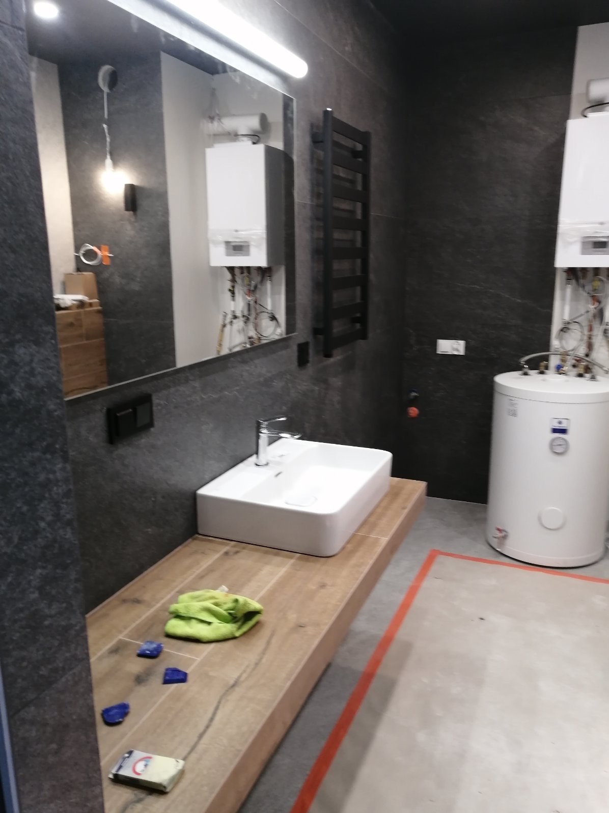 Nowoczesna łazienka w trakcie prac wykończeniowych: biała umywalka nablatowa, drewnopodobny blat, ciemne płytki na ścianach, widoczne instalacje grzewcze i akcesoria, zielona ściereczka...