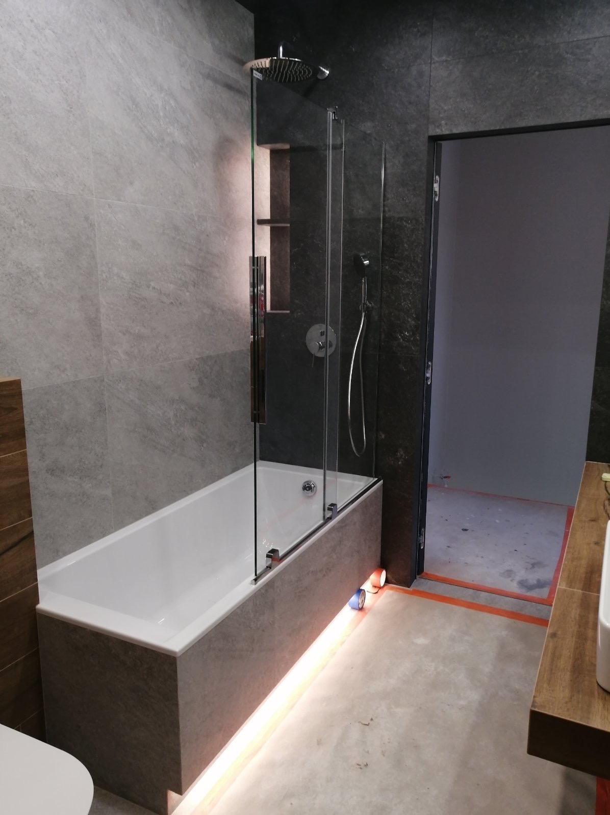 Nowoczesna łazienka z wanną obudowaną płytkami imitującymi beton, szklanym parawanem nawannowym i podświetleniem LED u dołu. Widoczna również deszczownica i fragment umywalki z blatem z drewna.