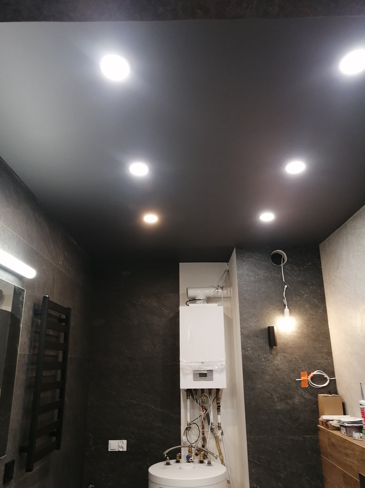 Ciemny sufit z oświetleniem punktowym w łazience z kamiennymi płytkami, widoczny bojler i grzejnik drabinkowy