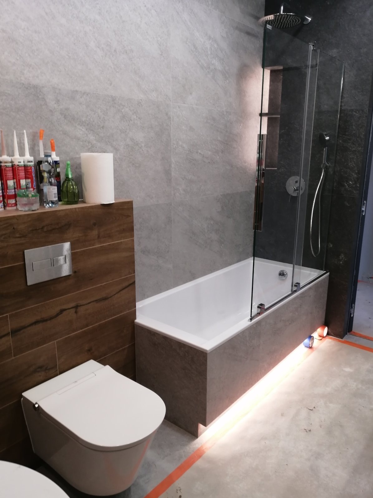 Nowoczesna łazienka z wanną obudowaną szarą płytką, szklanym parawanem nawannowym, podświetleniem LED i wiszącą toaletą z drewnopodobną zabudową ścienną. Widoczne silikony i akcesoria na półce.